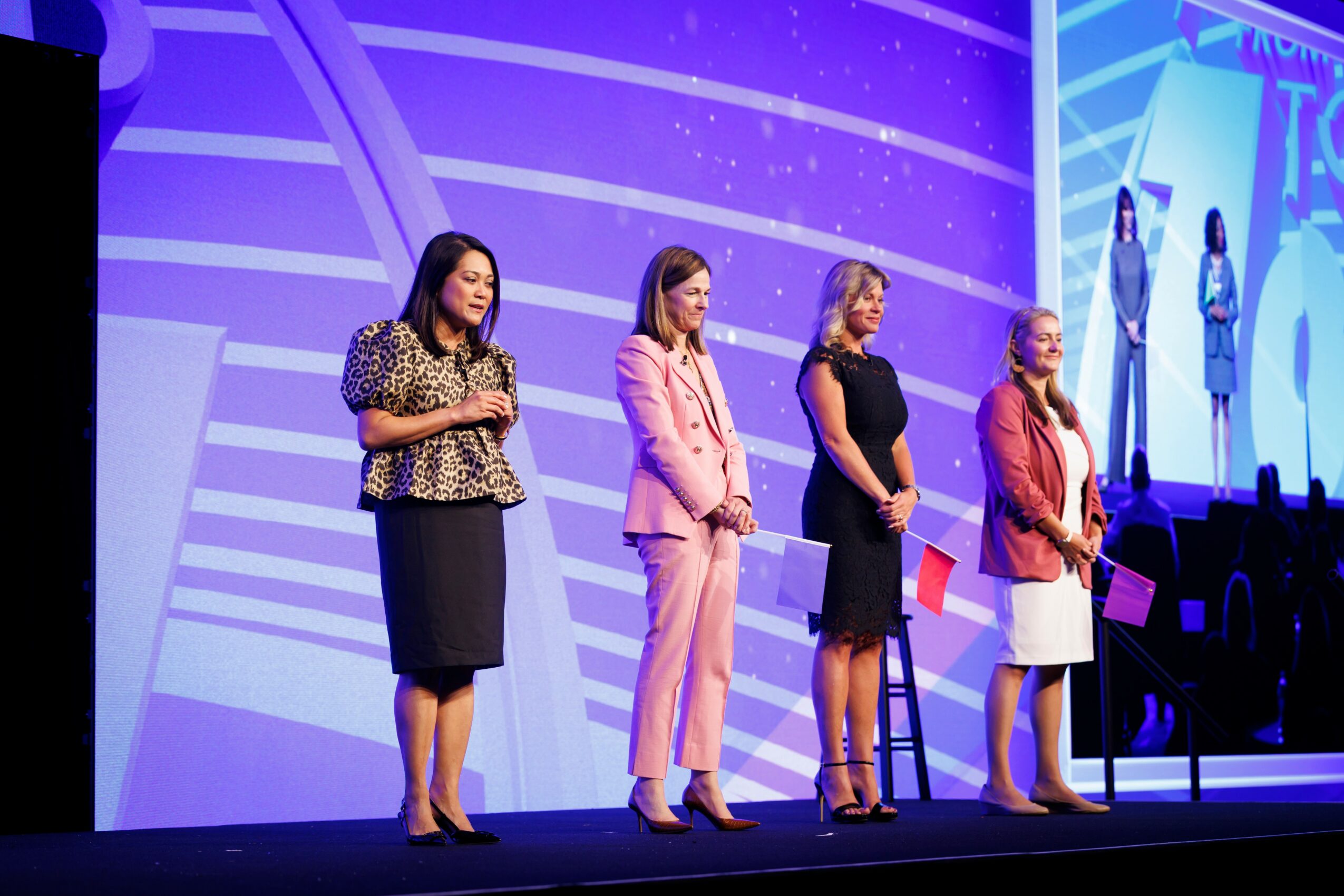 Four women stand on stage in front of a large screen, speaking into microphones during a presentation or event.