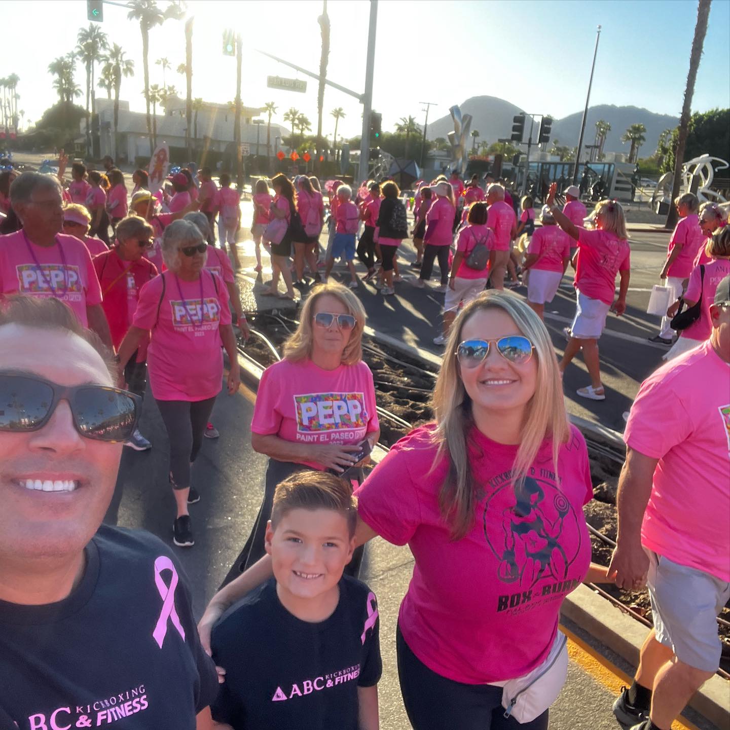 Group of people wearing pink shirts participating in an outdoor event, with palm trees and mountains in the background.