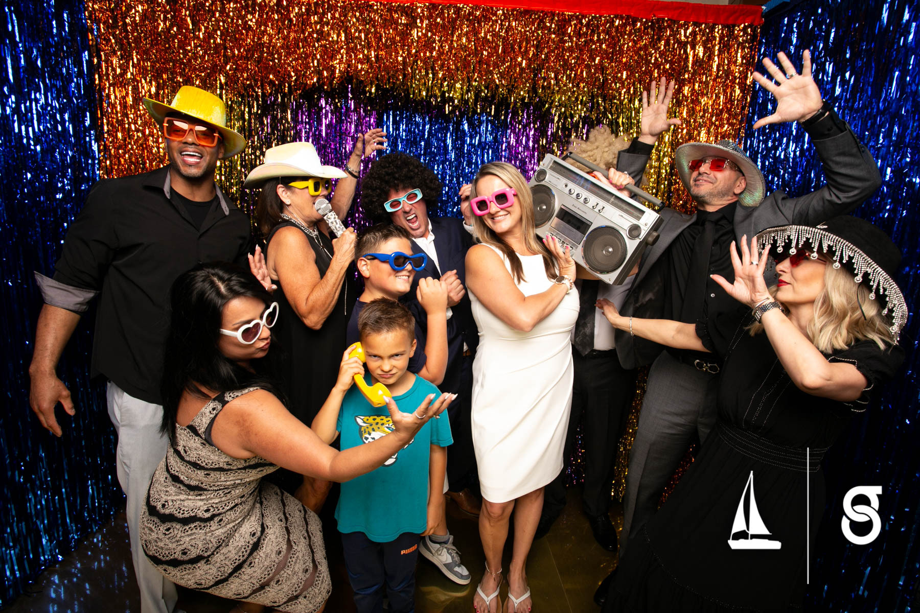 Group of people in costumes and accessories, some wearing hats and sunglasses, celebrating with a colorful backdrop.