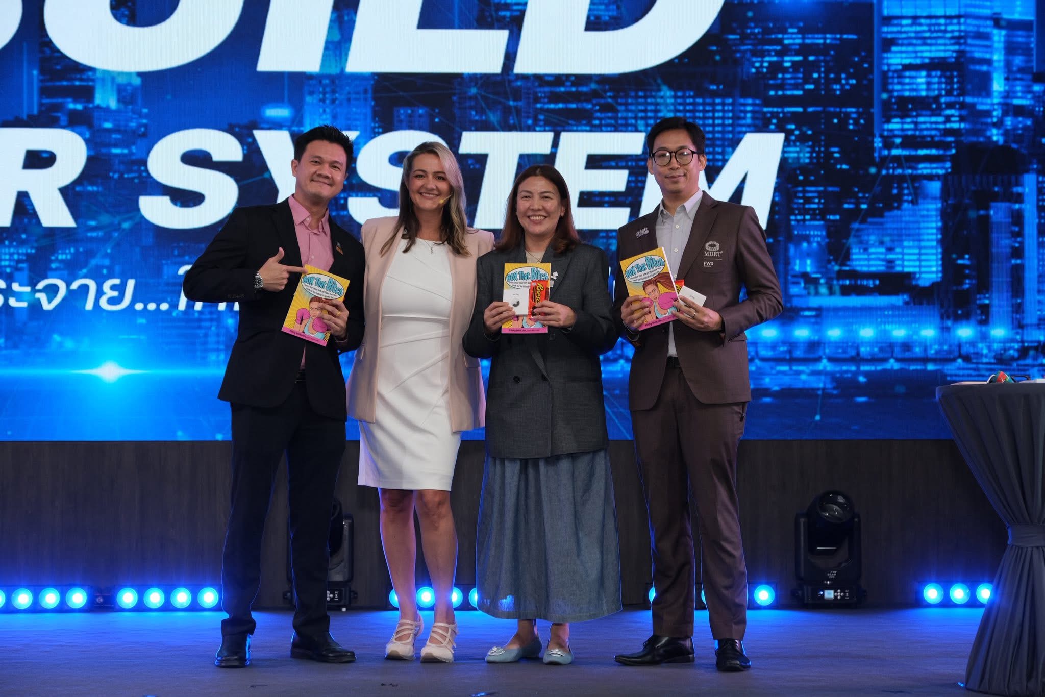 Four people standing on stage holding books, with a blue digital cityscape background and a large screen behind them.