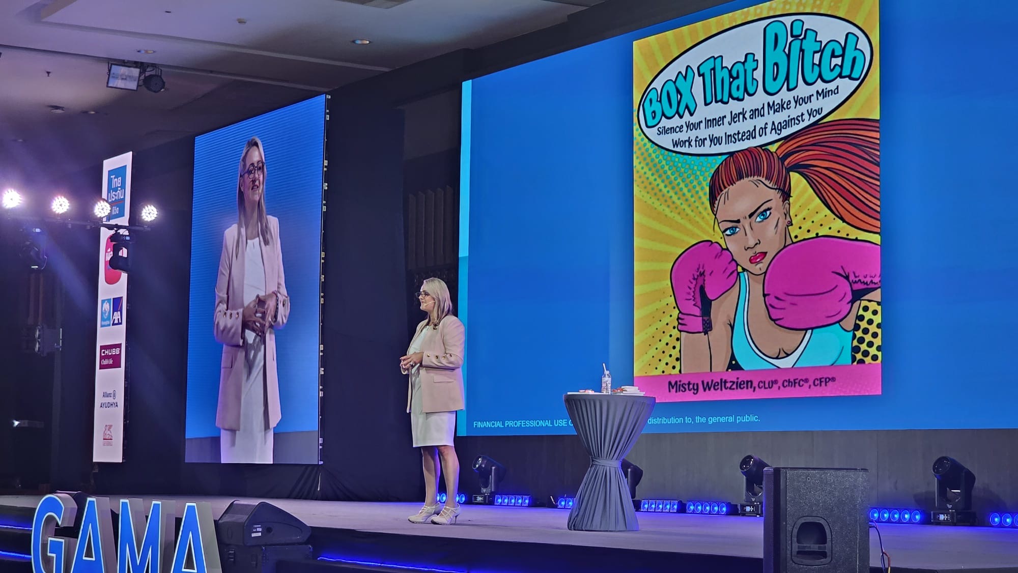 Woman speaking on stage with large screens behind her, one displaying a colorful cartoon book cover, in a conference setting.