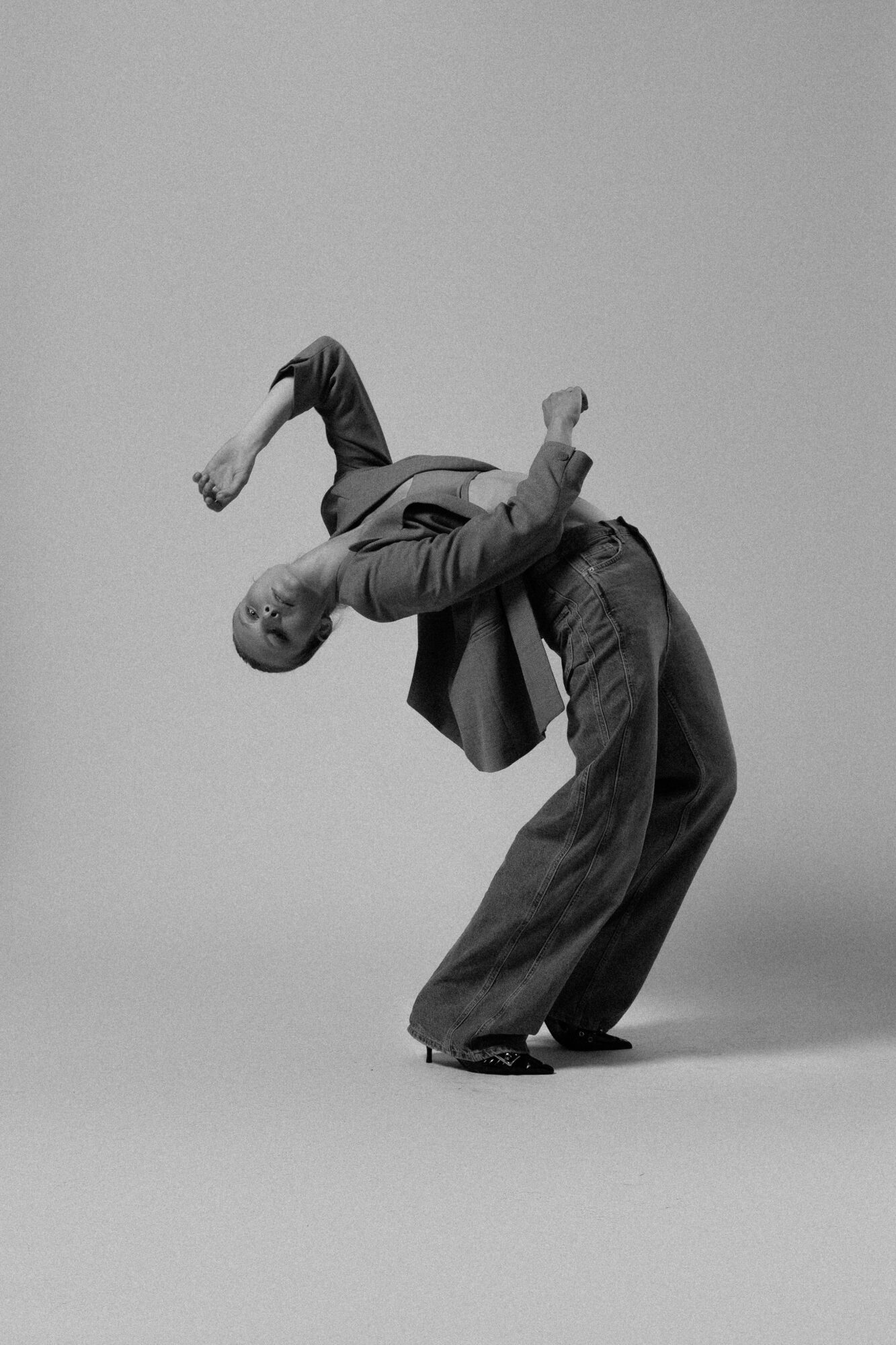 Person bending backward with arms raised, wearing a jacket and wide pants, against a plain background.