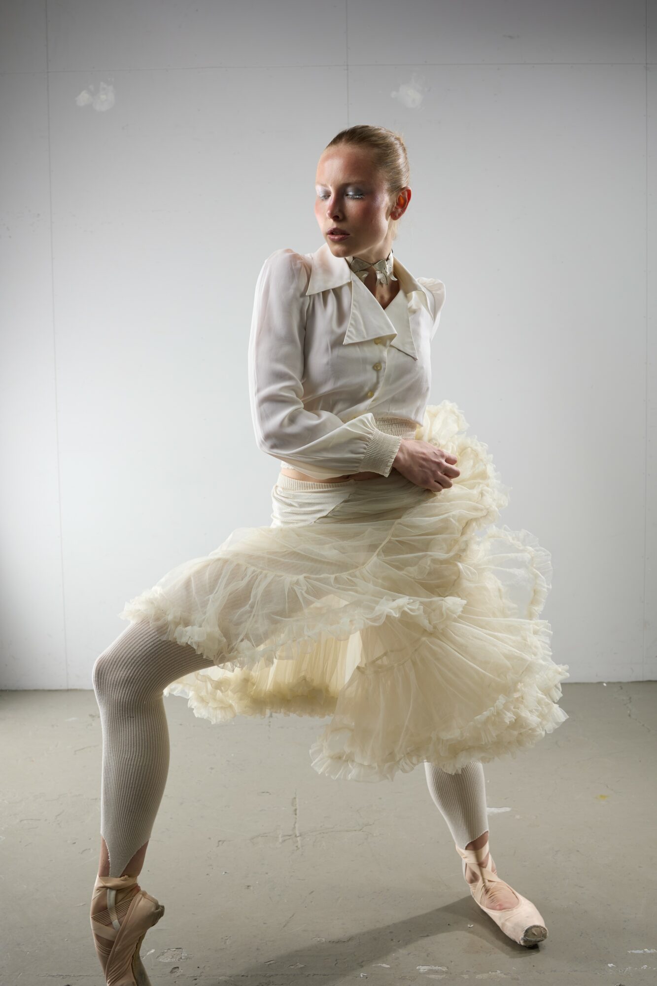 A person in a ballet costume poses with one leg raised and arms crossed, against a plain background.