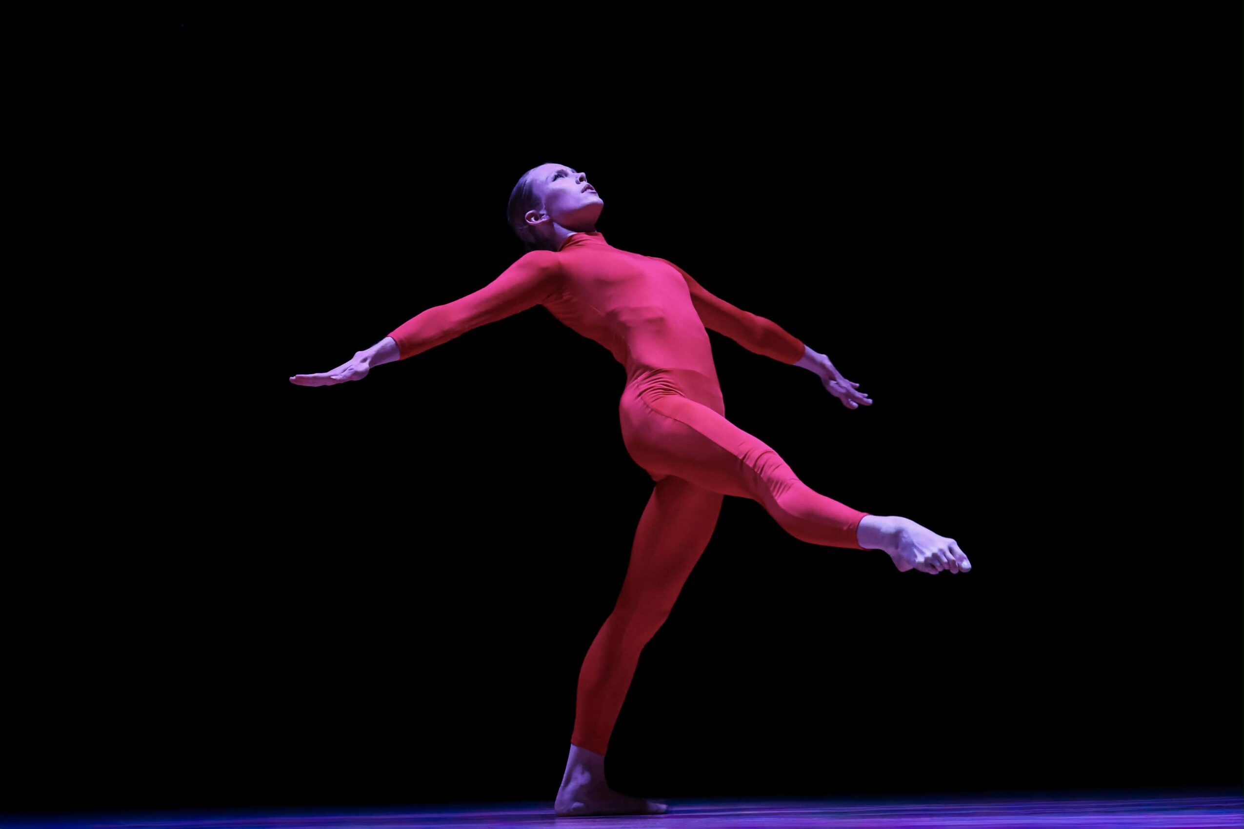 A person in a pink outfit performs a dance move with arms and leg extended, against a black background.