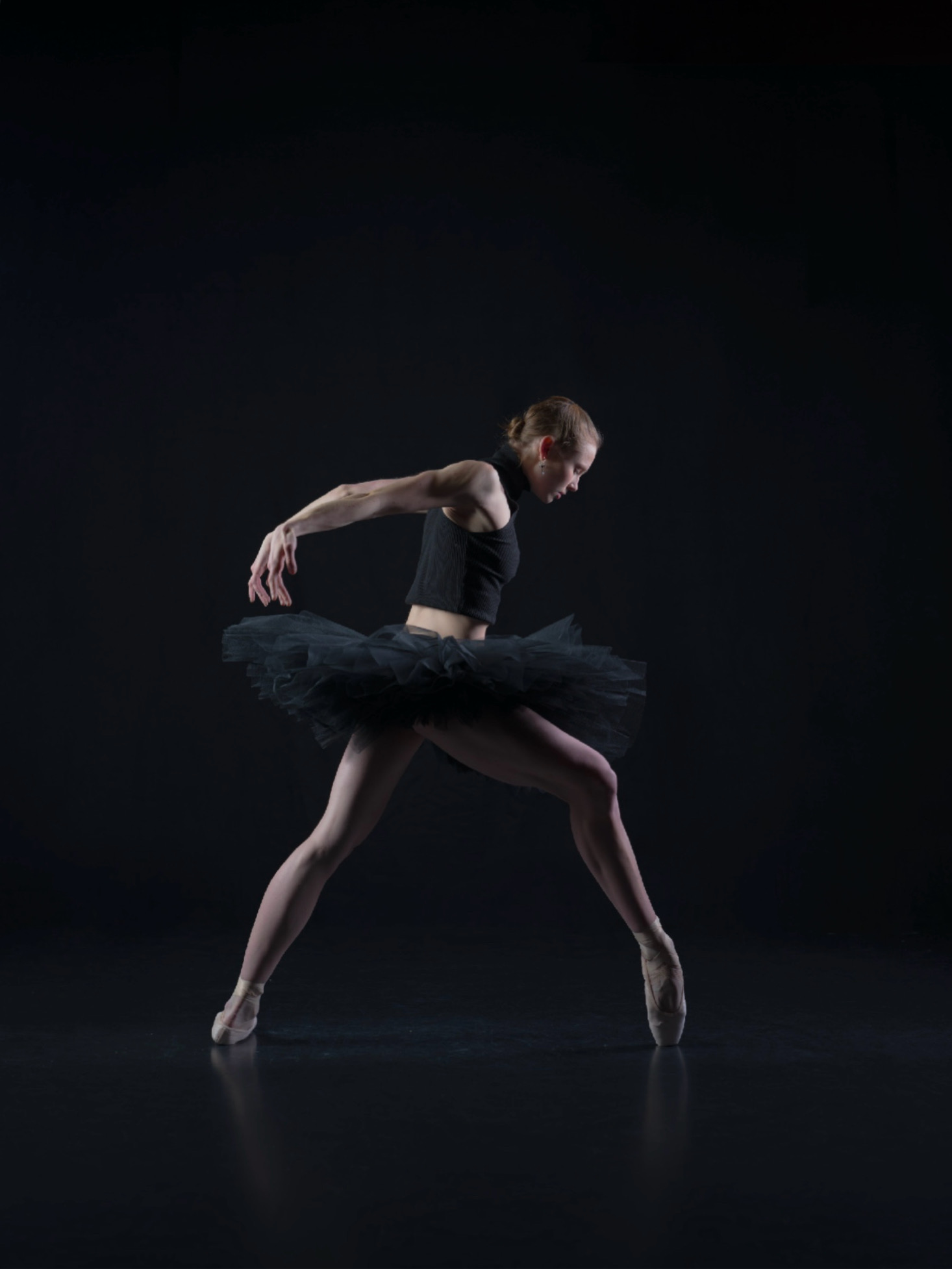 Ballet dancer in black tutu and tights performs on dark stage, one leg extended, arms curved gracefully.