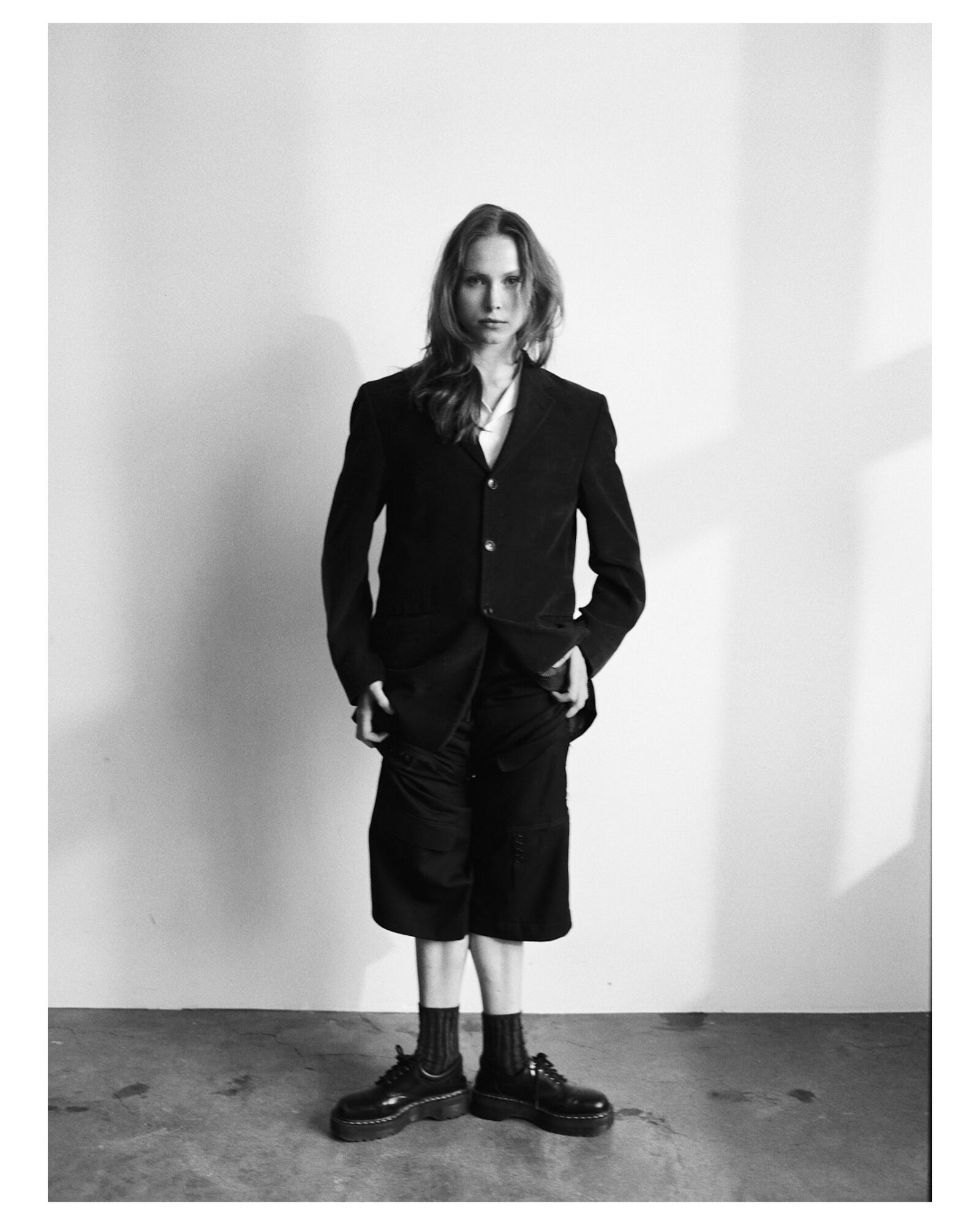 Person with long hair wearing a dark blazer, shorts, and boots standing against a plain wall.