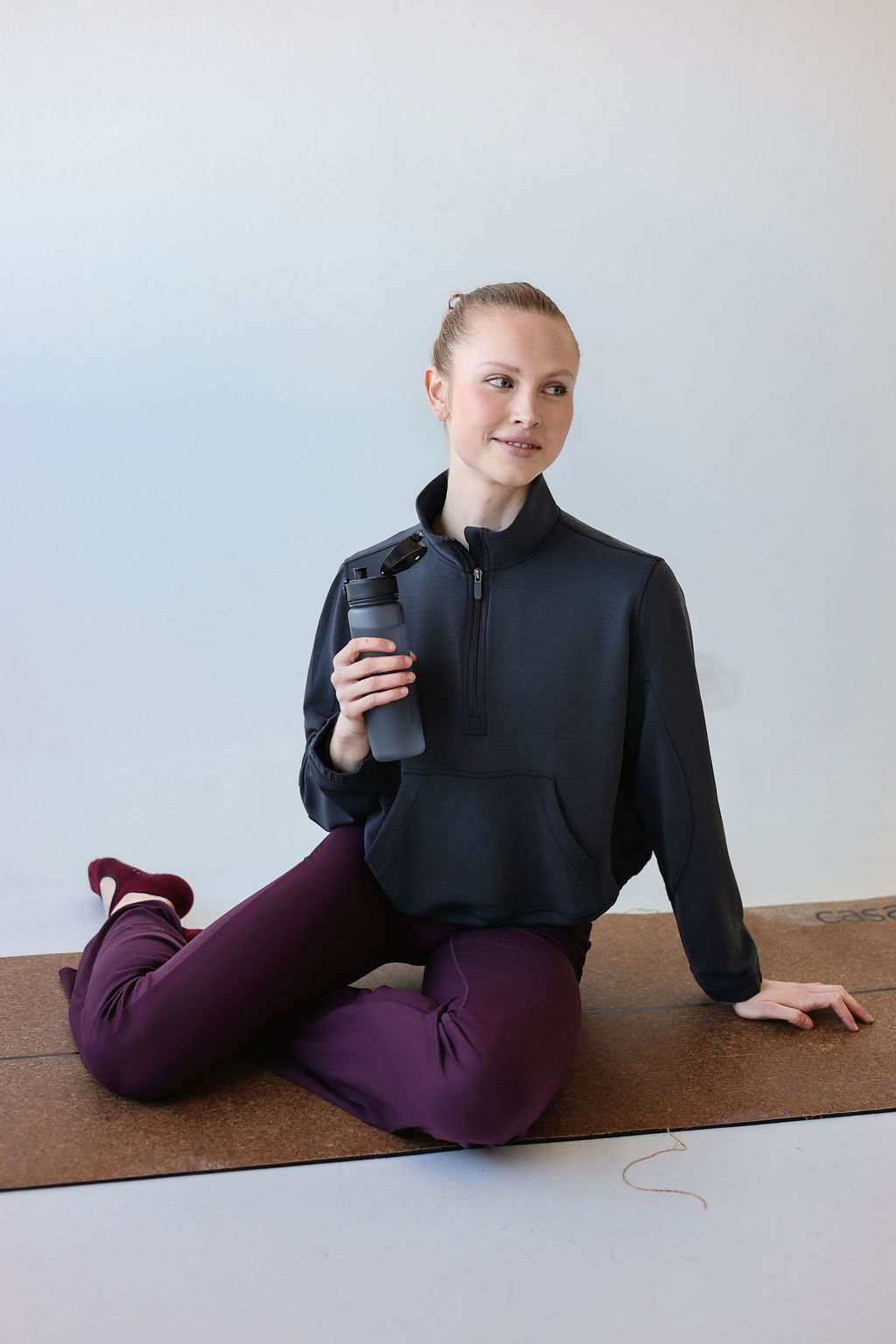 Woman sitting on yoga mat holding a water bottle, wearing a black jacket and purple pants, against a plain background.