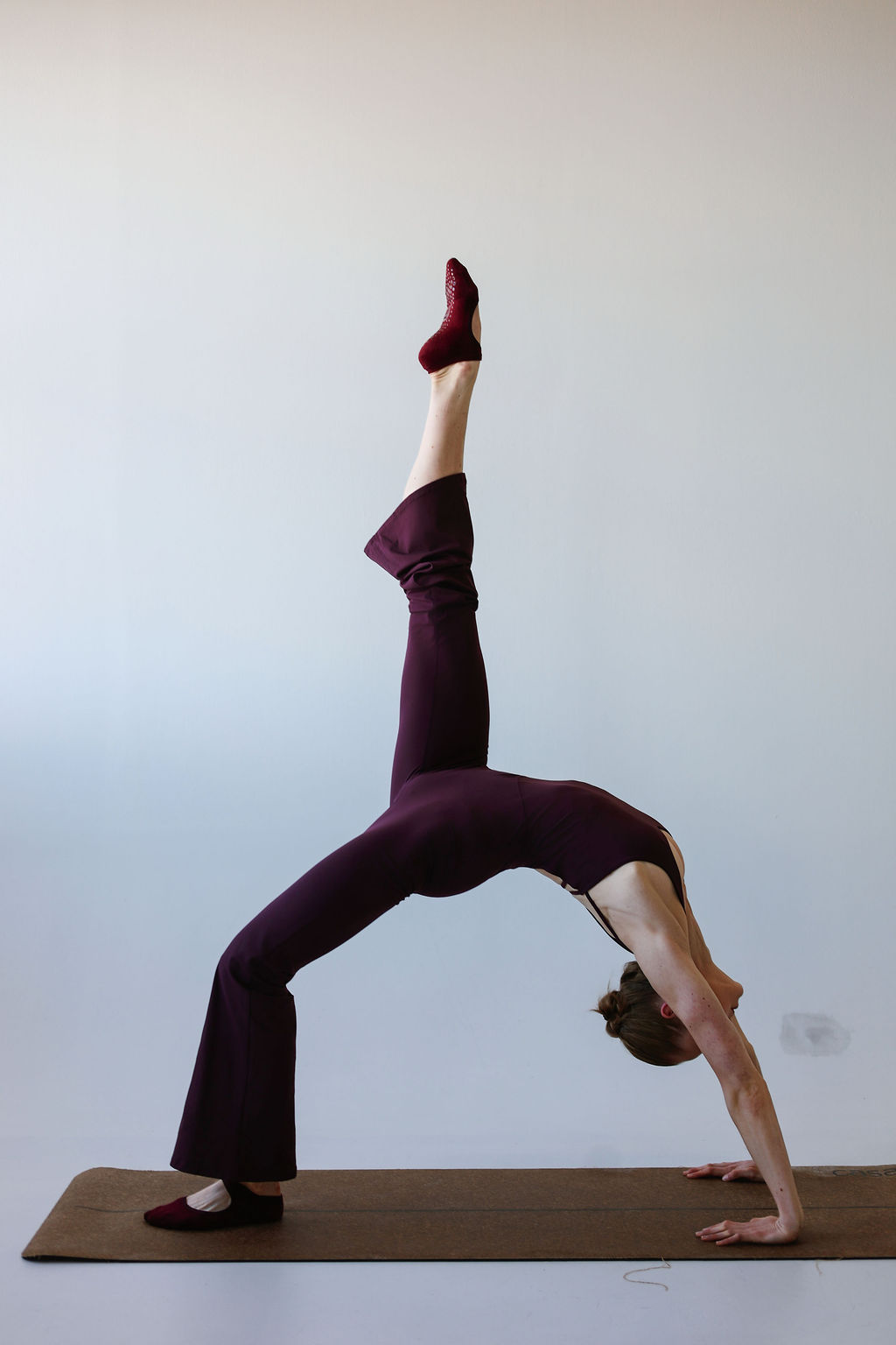 Person performing a backbend yoga pose with legs extended upward, head and hands on the floor, on a mat against a plain wall.