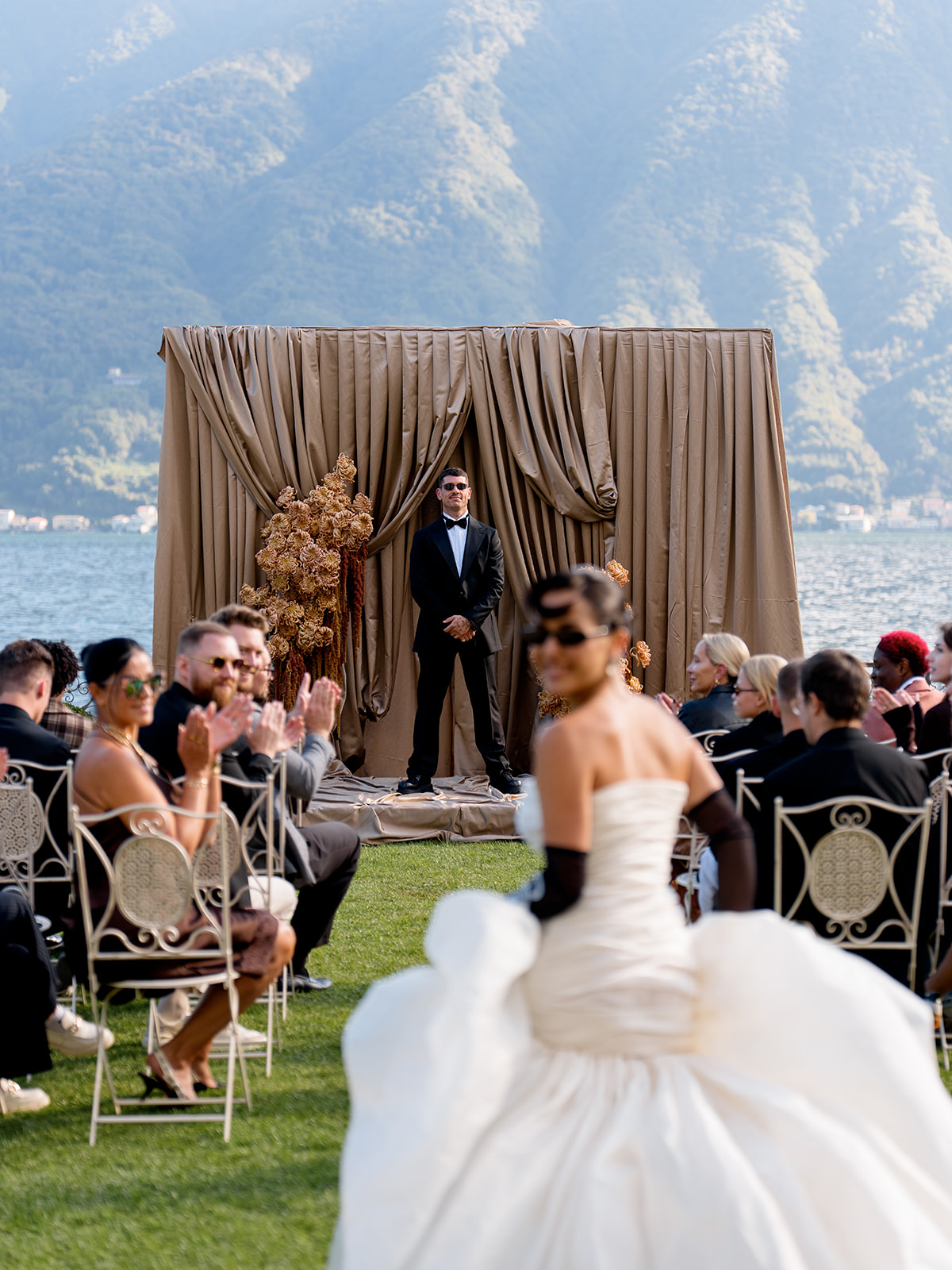 Bride in white wedding dress walking towards a man in a tuxedo on a stage outdoors, with seated guests and a lake backdrop.