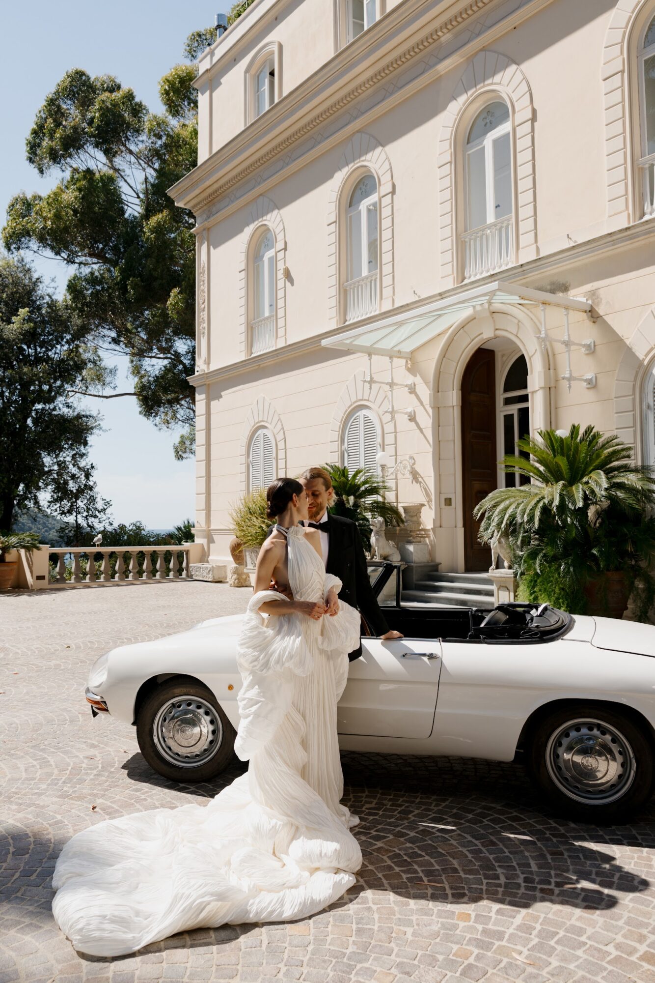 Bride in wedding dress standing next to a white convertible car outside a large white building with arched windows and greenery