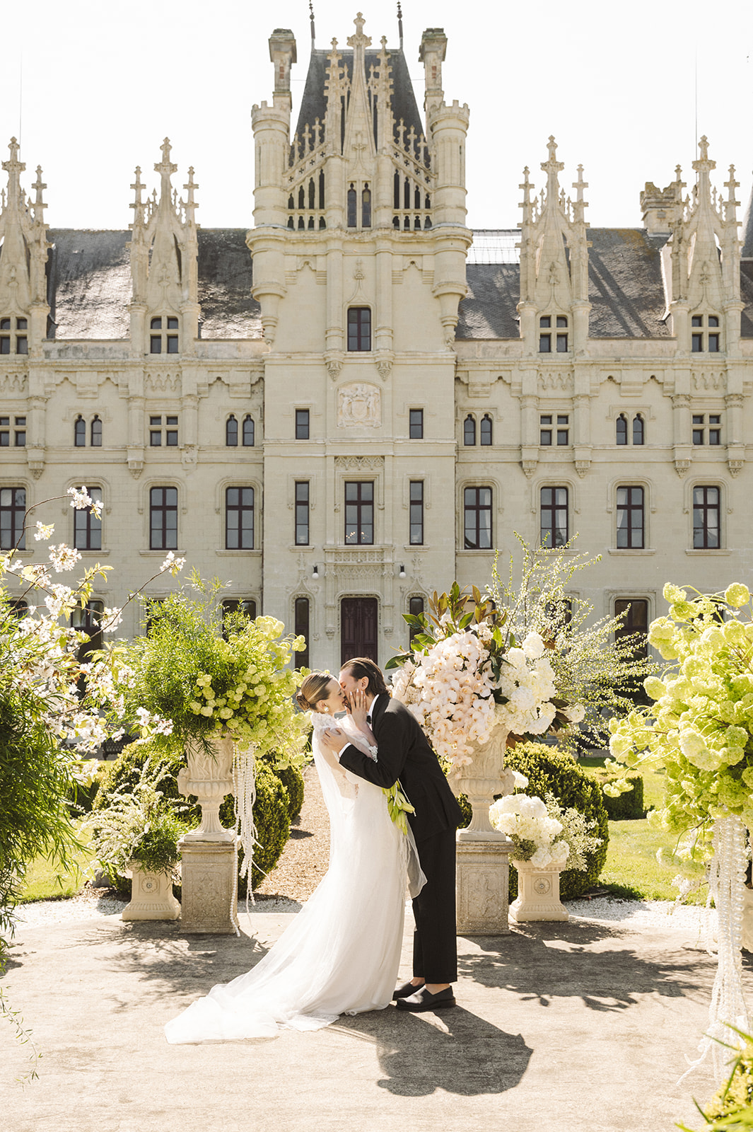 Couple in wedding attire kissing outdoors in front of a castle with floral arrangements and greenery.