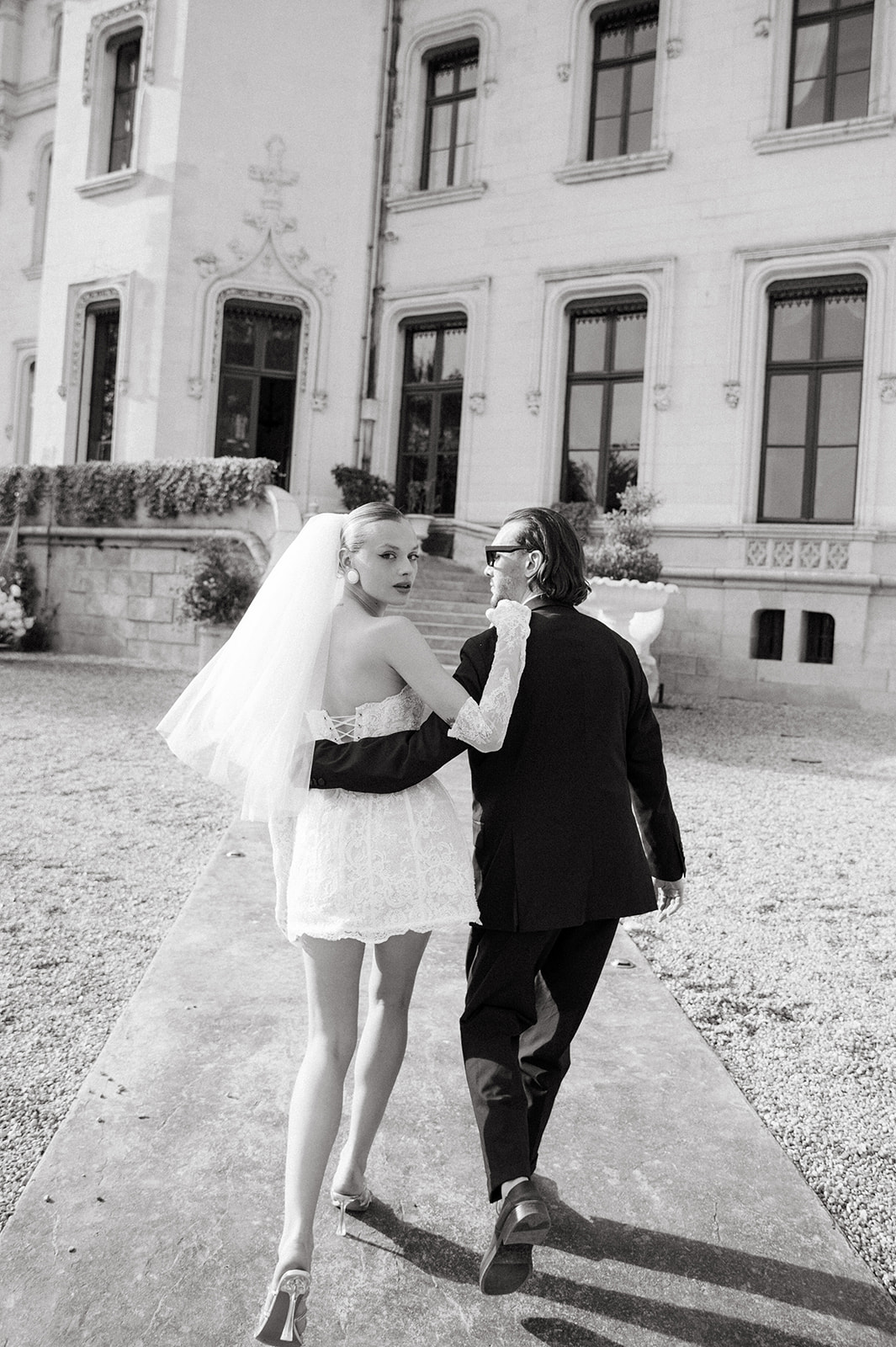 Two women walk together outside a historic building, one in a wedding dress and veil, the other in a suit.