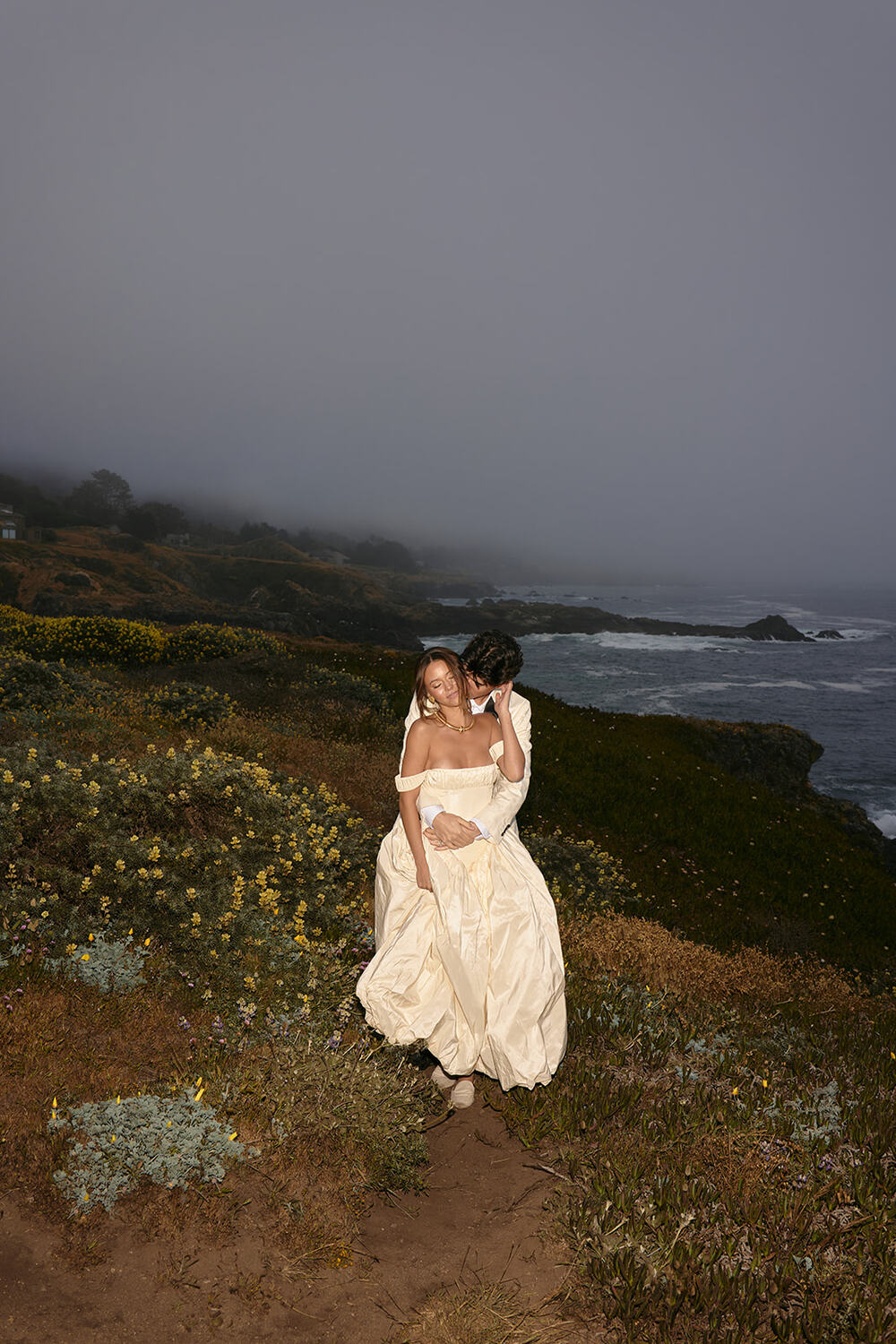 Two people in wedding attire standing on a grassy coastal landscape with foggy sky and ocean in background.