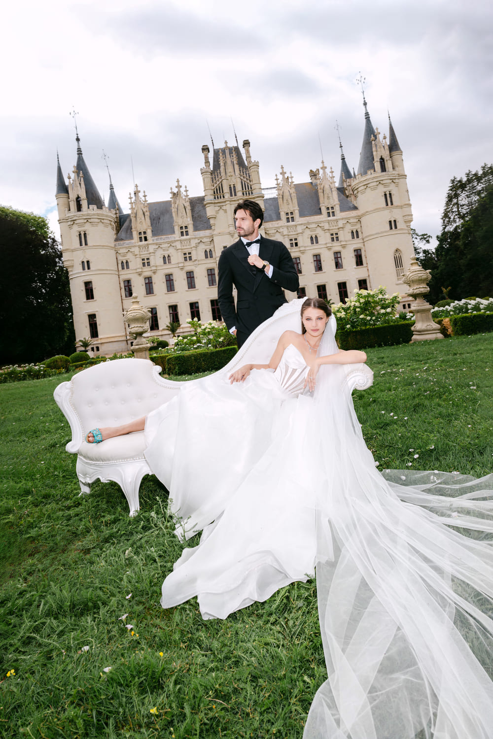 Bride in a white wedding gown reclining on a white sofa outdoors, groom in a suit standing behind her, castle in background