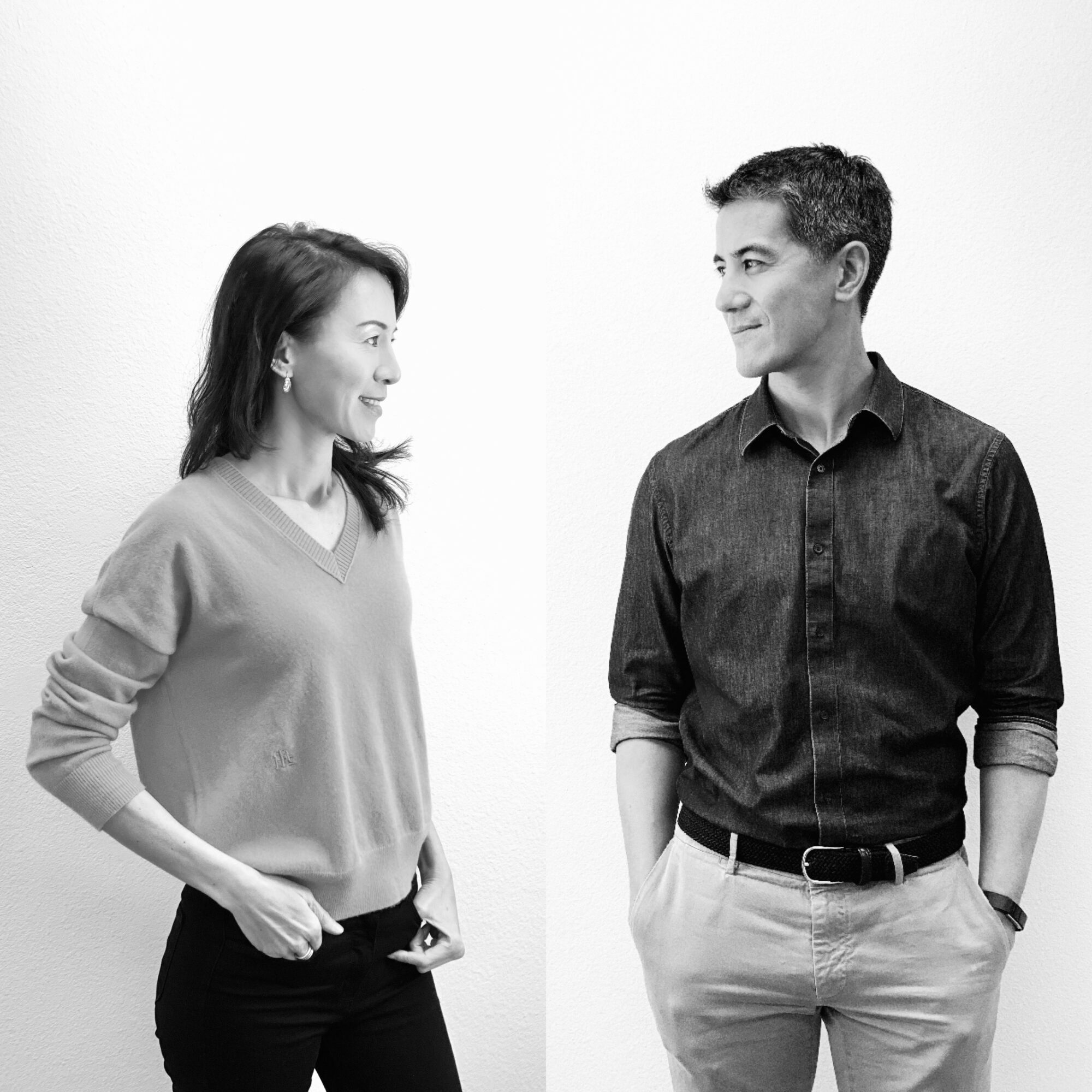 Two people, a woman and a man, face each other and smile, standing against a plain white background.