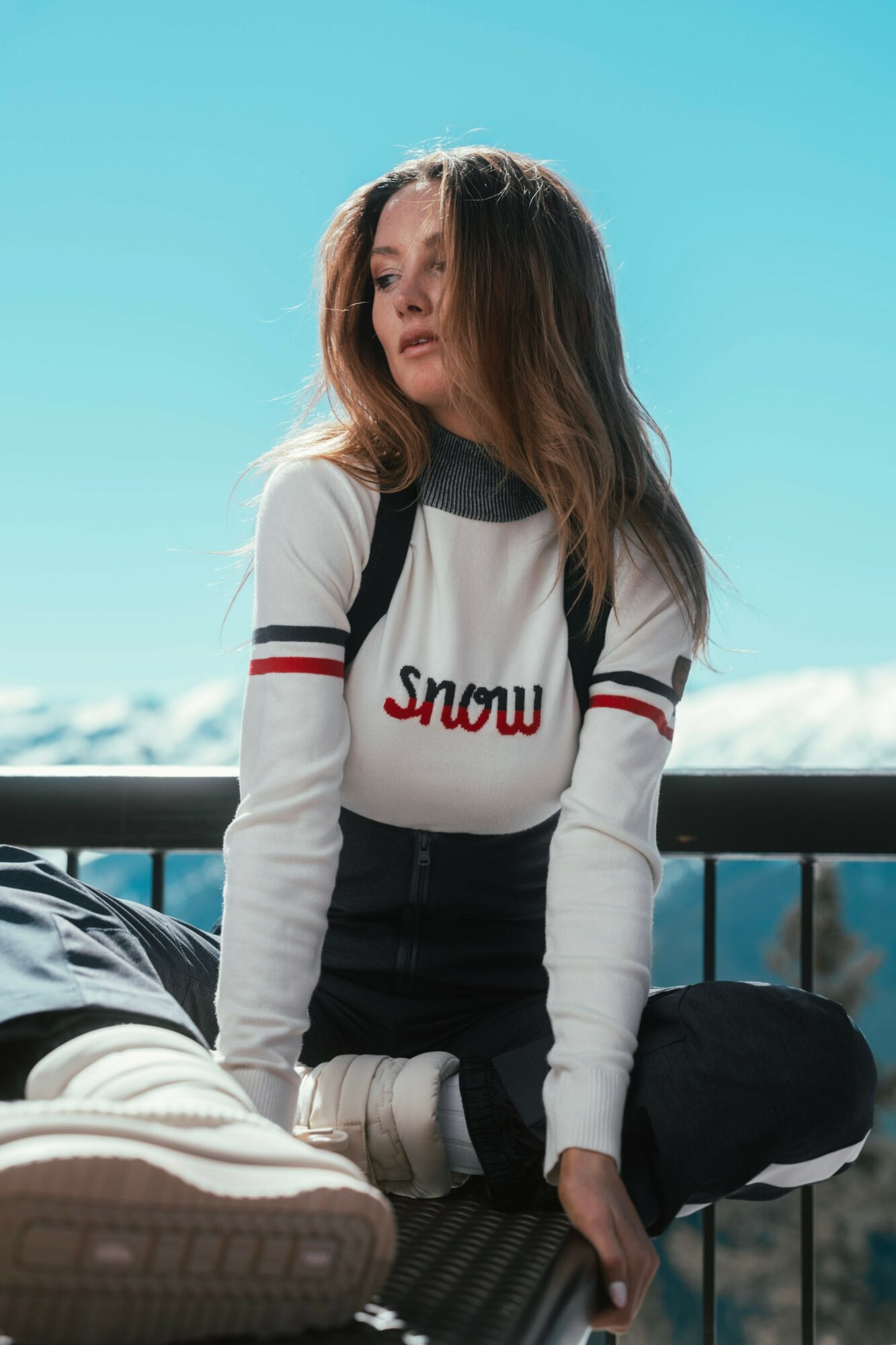 Young woman with long hair wearing a white sweater with red and black stripes, sitting outdoors with a blue sky background.