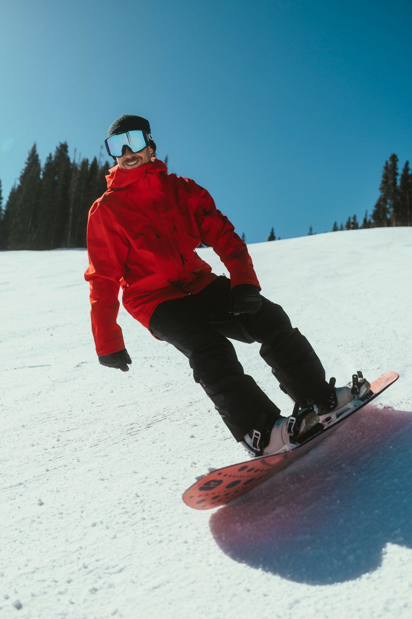 Person snowboarding on snow-covered slope wearing red jacket, black pants, goggles, and helmet, with trees in background.