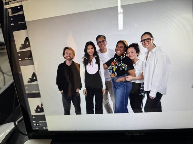 Group of six diverse people standing together indoors, smiling, with plain white wall background.