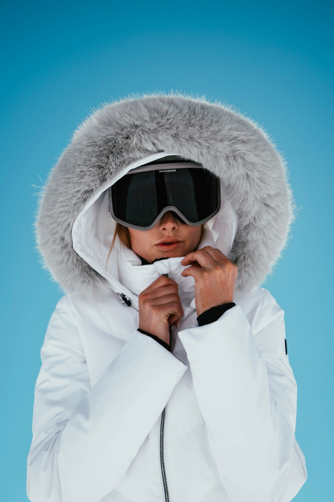 Child in winter clothing with fur-lined hood and goggles, holding the hood's drawstrings, against a blue background.