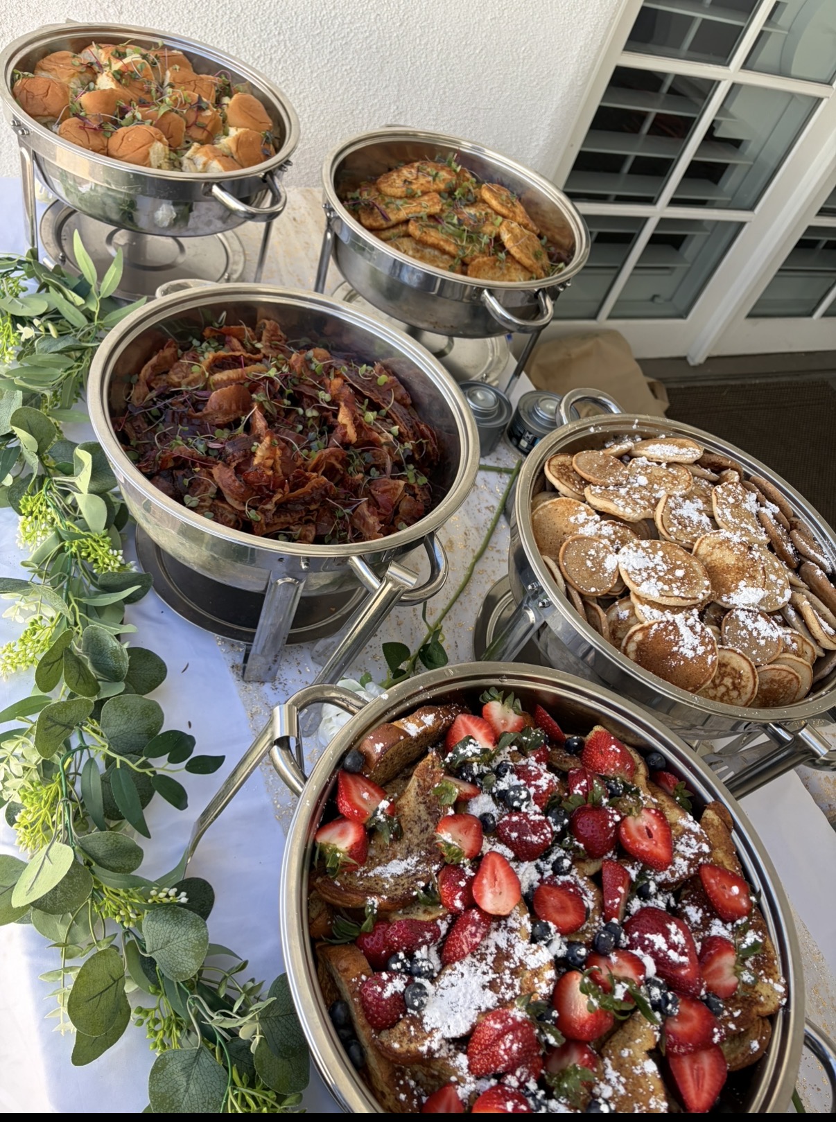 Five bowls of desserts and dishes on a table with greenery, viewed from above, with a window in the background.