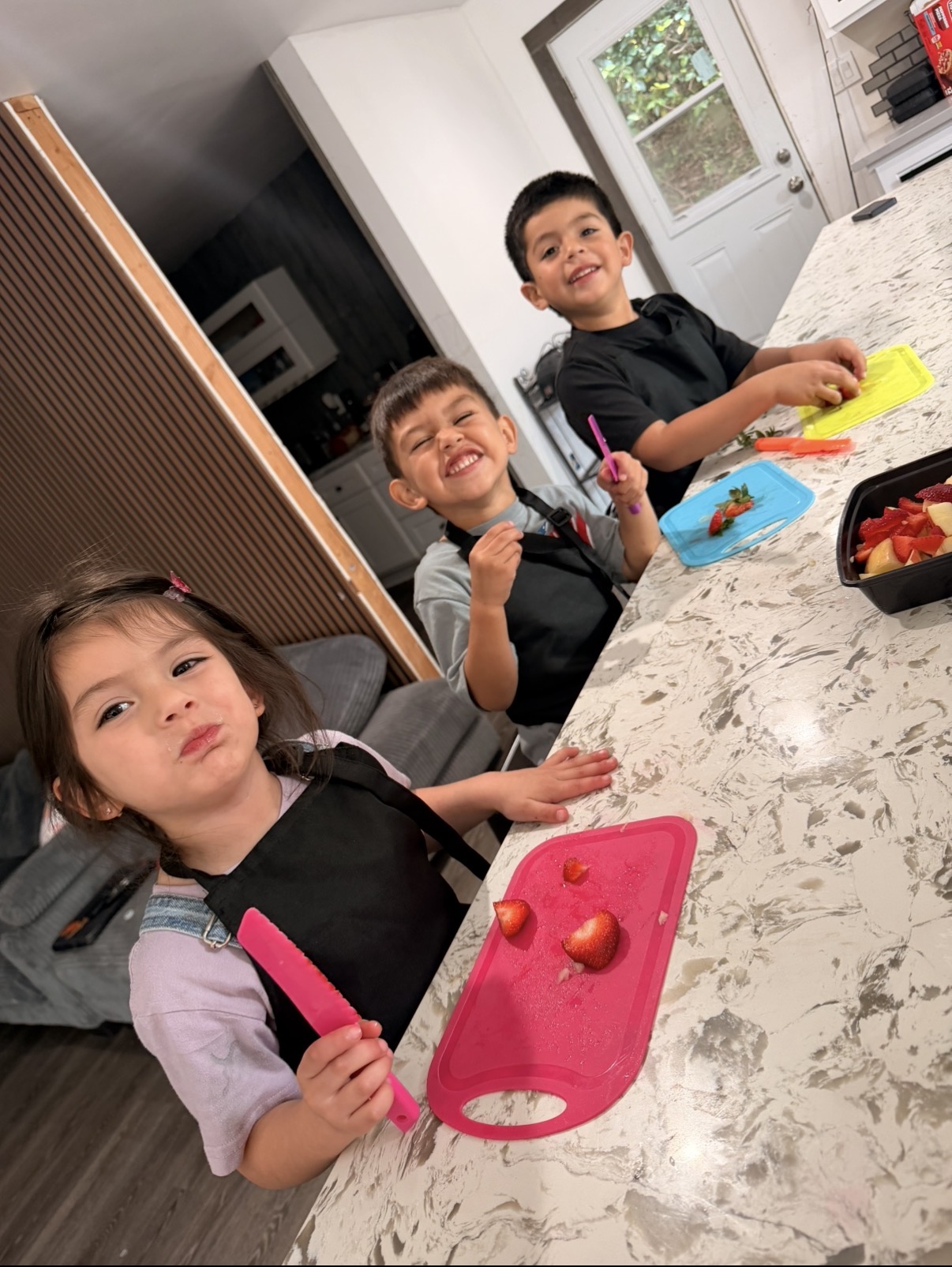 Three children sitting at a kitchen counter with colorful cutting boards and food, smiling and preparing snacks.
