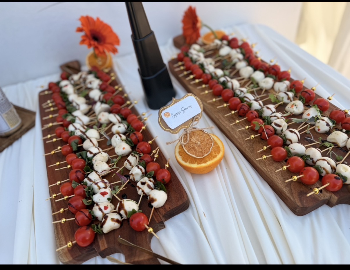 Two wooden boards with skewered appetizers, a small orange slice, and a sign, on a white tablecloth.