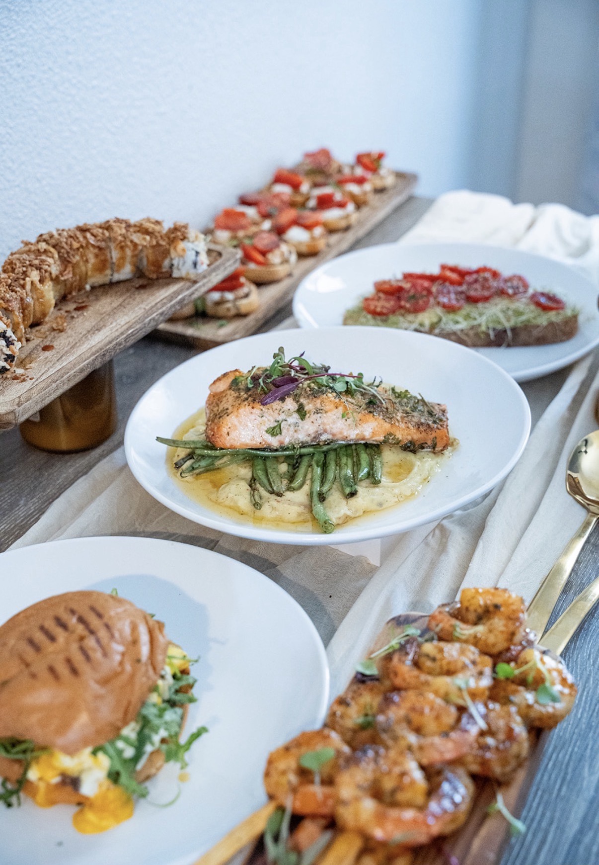 Assorted dishes including a fish fillet with green vegetables, a burger, and a dessert with strawberries on a table.