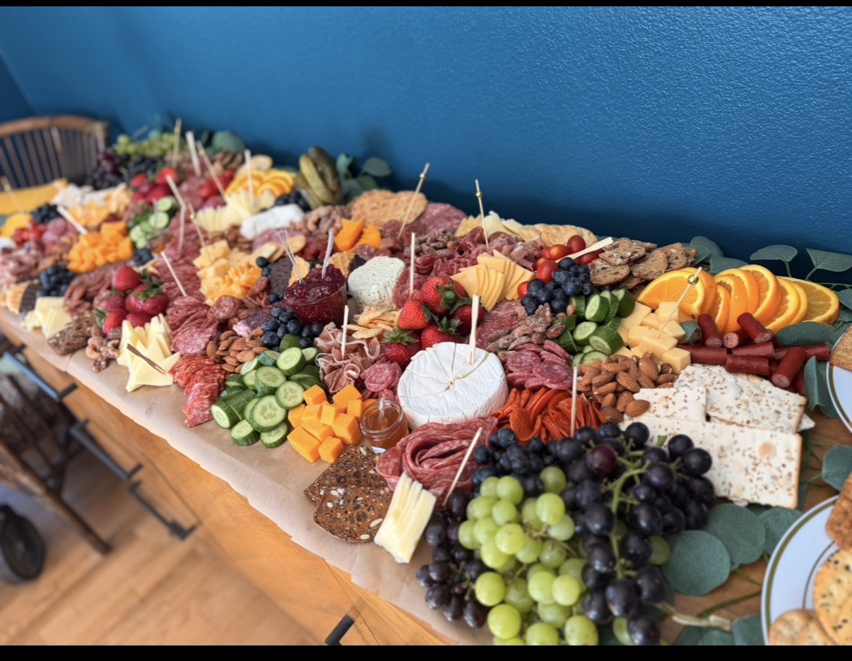 A long table with various cheeses, fruits, and snacks arranged on it, against a blue wall.