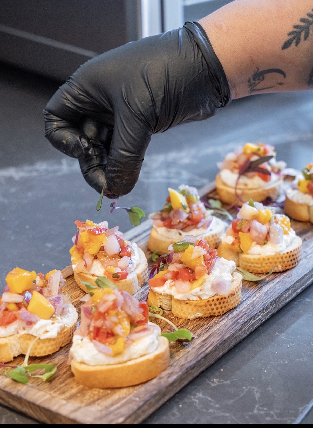 Hand wearing a black glove garnishes small appetizer bites on a wooden board with a green herb, with toppings including diced fruit and sauce.