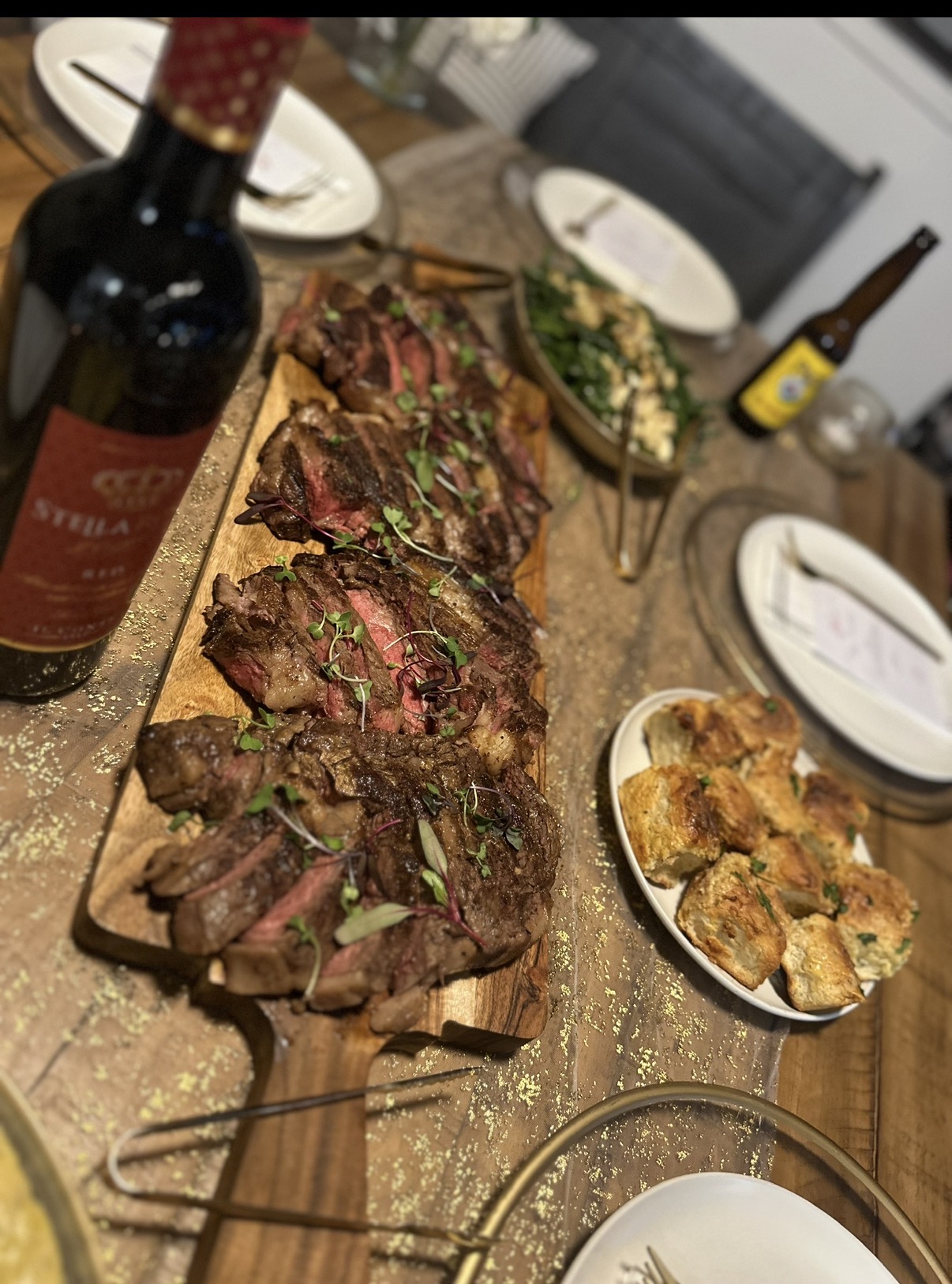 Sliced grilled steak on a wooden board with green garnish, surrounded by plates, a bottle, and a bowl of breaded appetizers on a table.