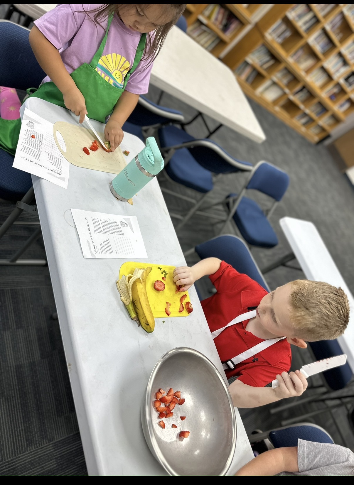 Children at a table with craft supplies, including a yellow toy, a bowl with small objects, and paper, in a classroom.
