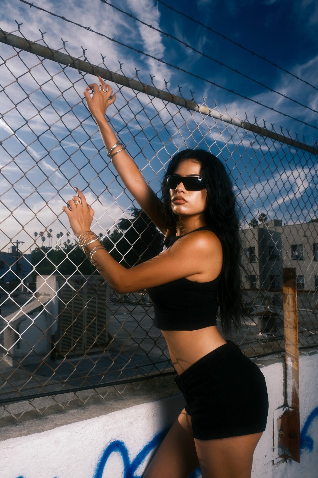 Woman with long dark hair wearing sunglasses, black crop top, and shorts, standing by a chain-link fence outdoors.