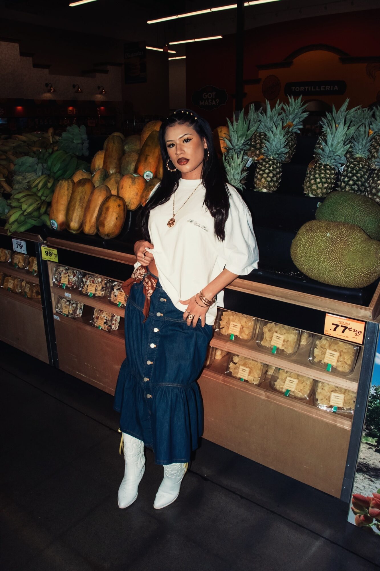 Woman with dark hair and red lipstick stands in a grocery store aisle with pineapples and melons behind her.
