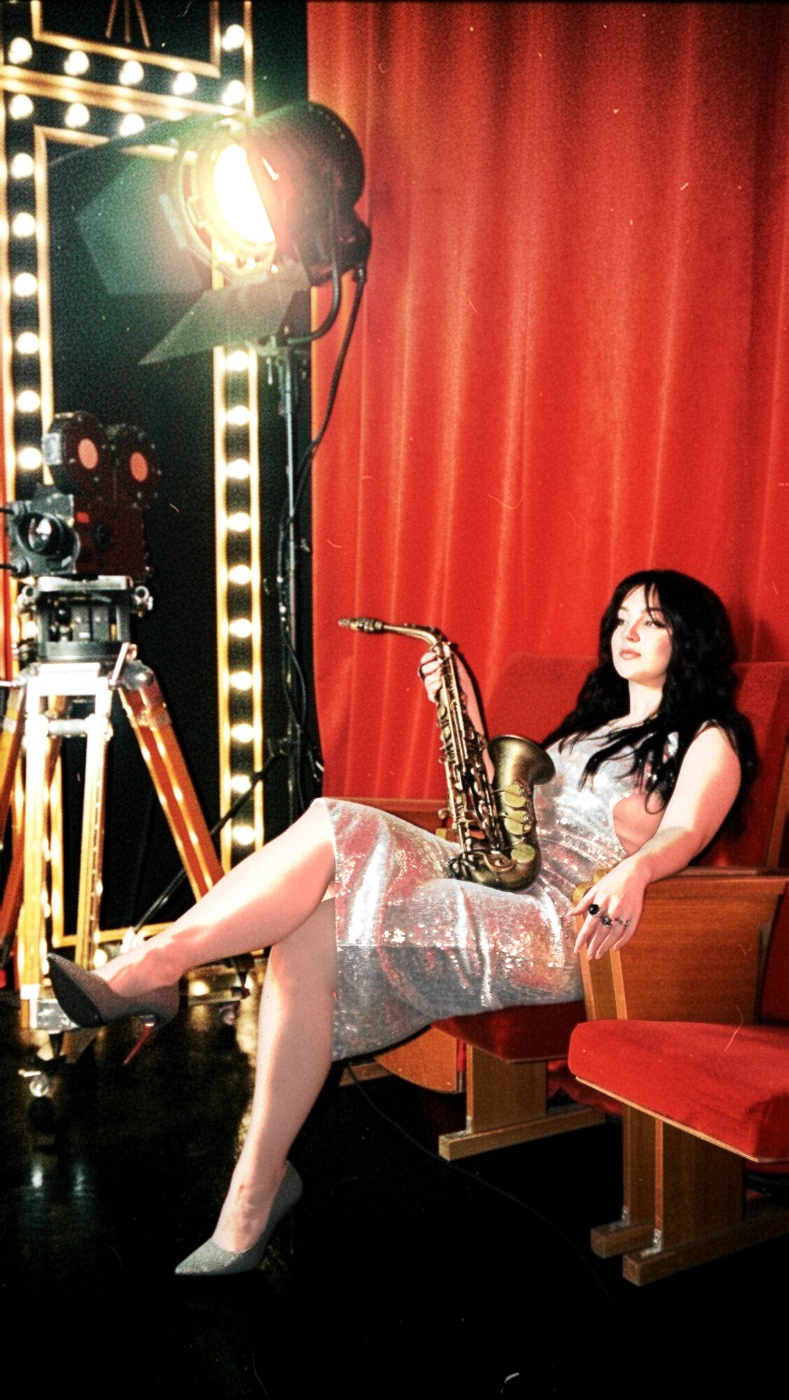 Woman sitting on a bench holding a saxophone, with stage lights and red curtain background.