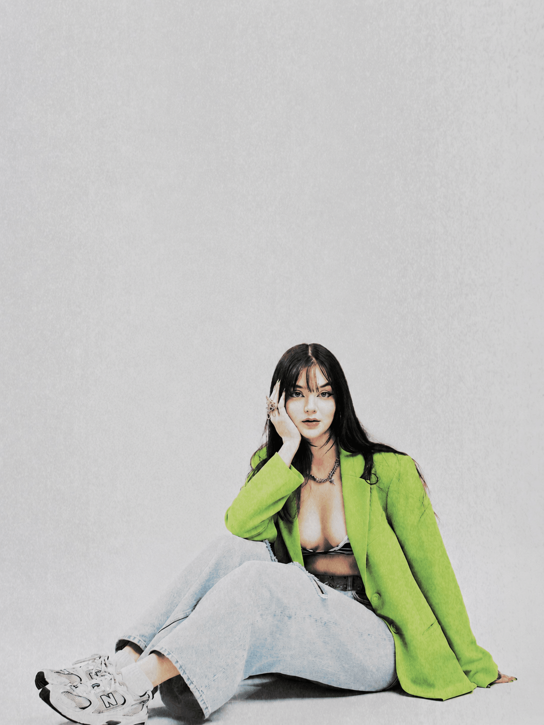 Woman sitting on the floor with a gray background, wearing a bright green jacket and light-colored pants.