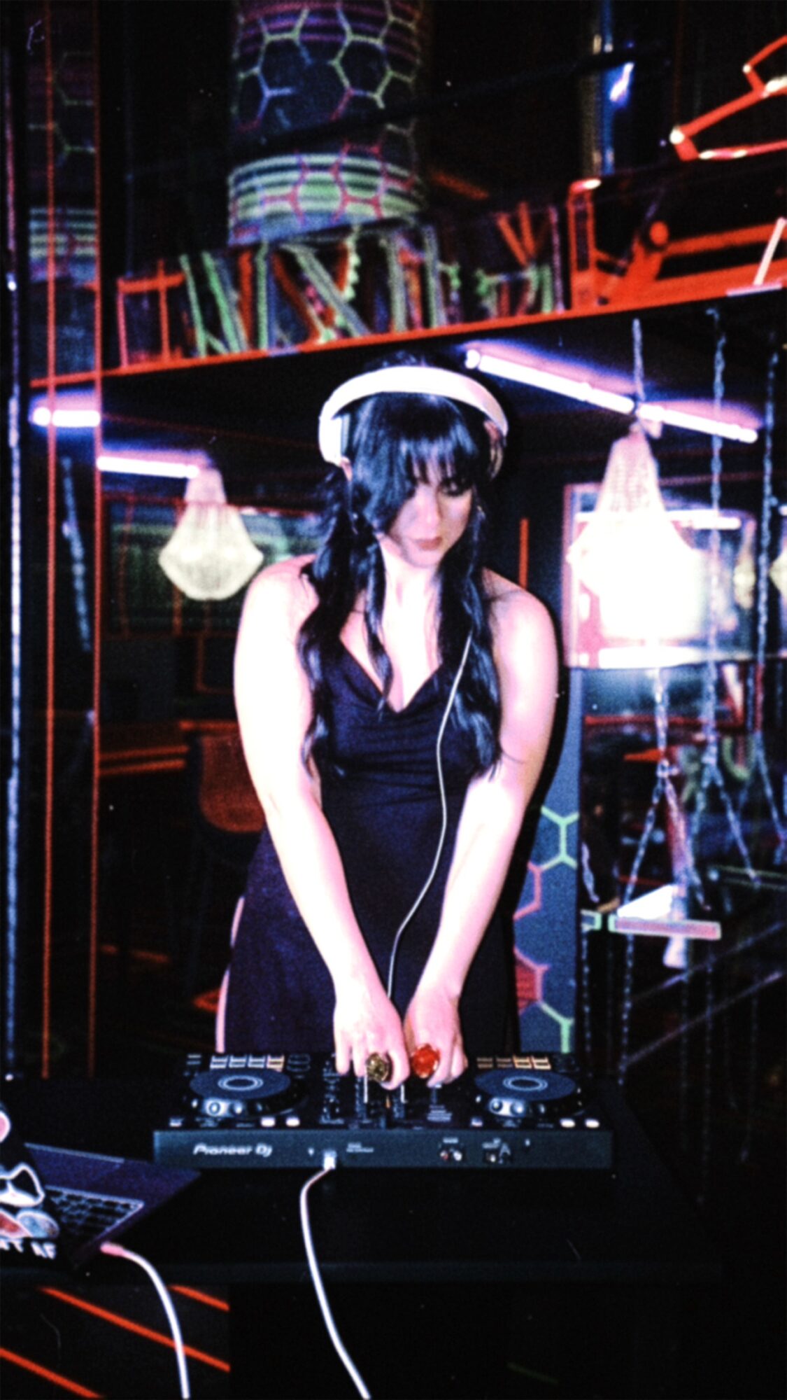 Young woman with long dark hair wearing headphones and a black dress, DJing in a neon-lit club setting.