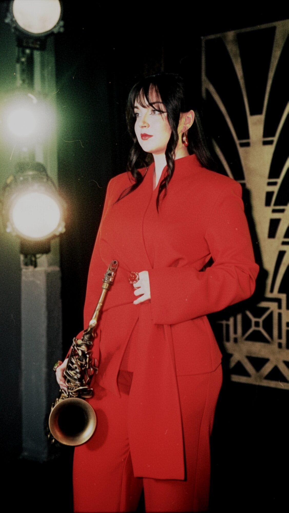 Woman in red coat holding a saxophone, standing in front of stage lights and decorative backdrop.