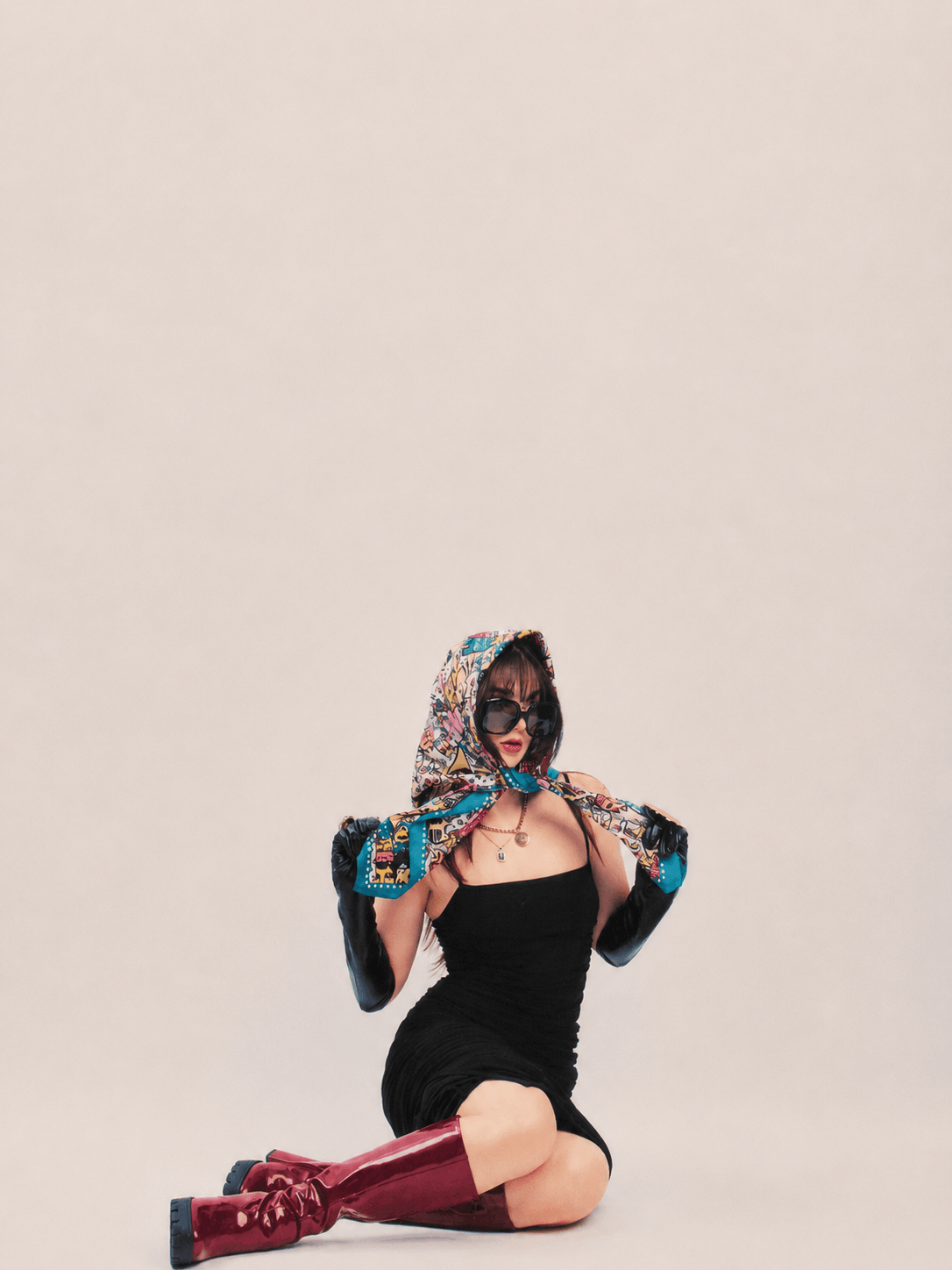 Person sitting on the floor wearing a black dress, sunglasses, and a colorful headscarf, holding the scarf with both hands.