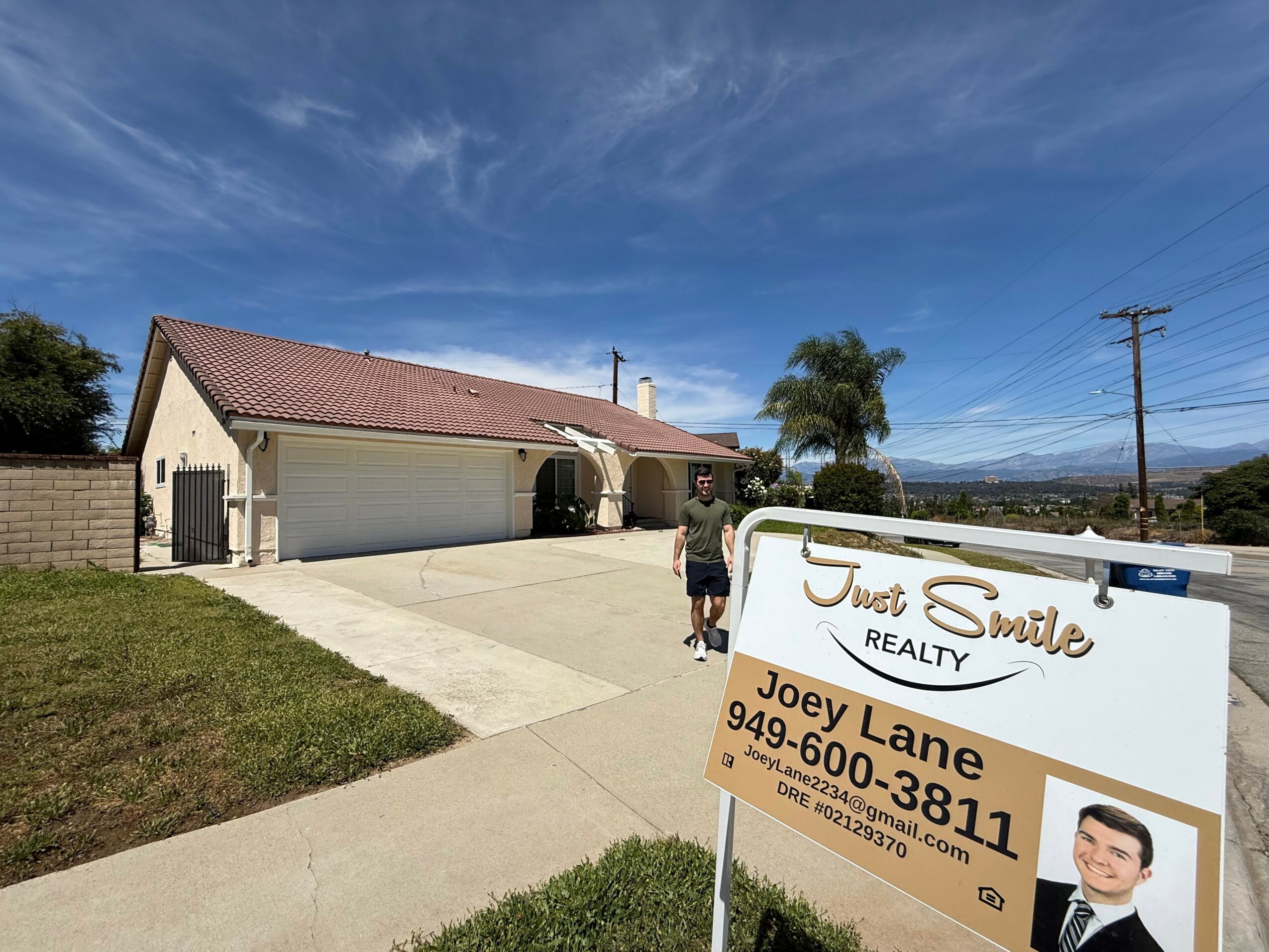 A house with a driveway, a person standing near a sign, and a clear blue sky with some clouds.