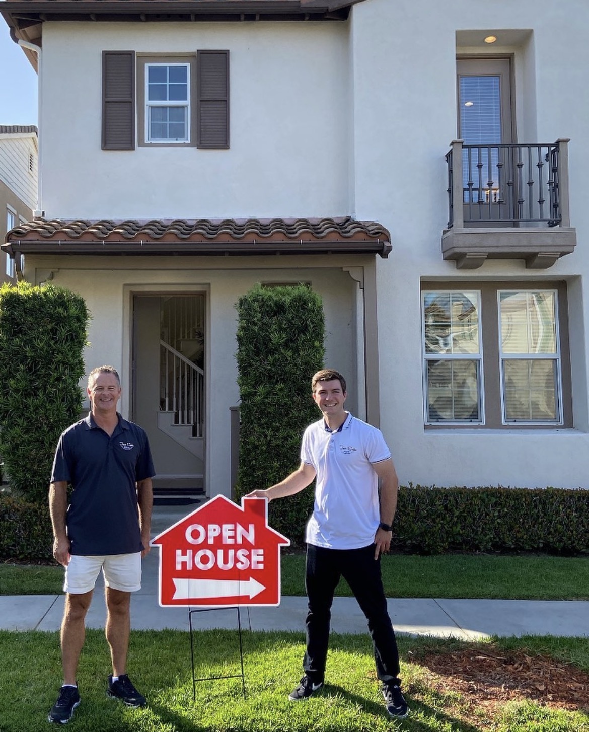Two men standing outside a house holding an open house sign, with a lawn and house in the background.