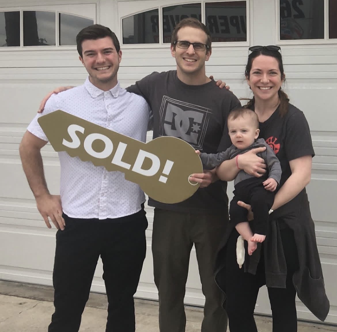 Group of three adults and a baby standing in front of a garage door, holding a 'SOLD!' sign, smiling.