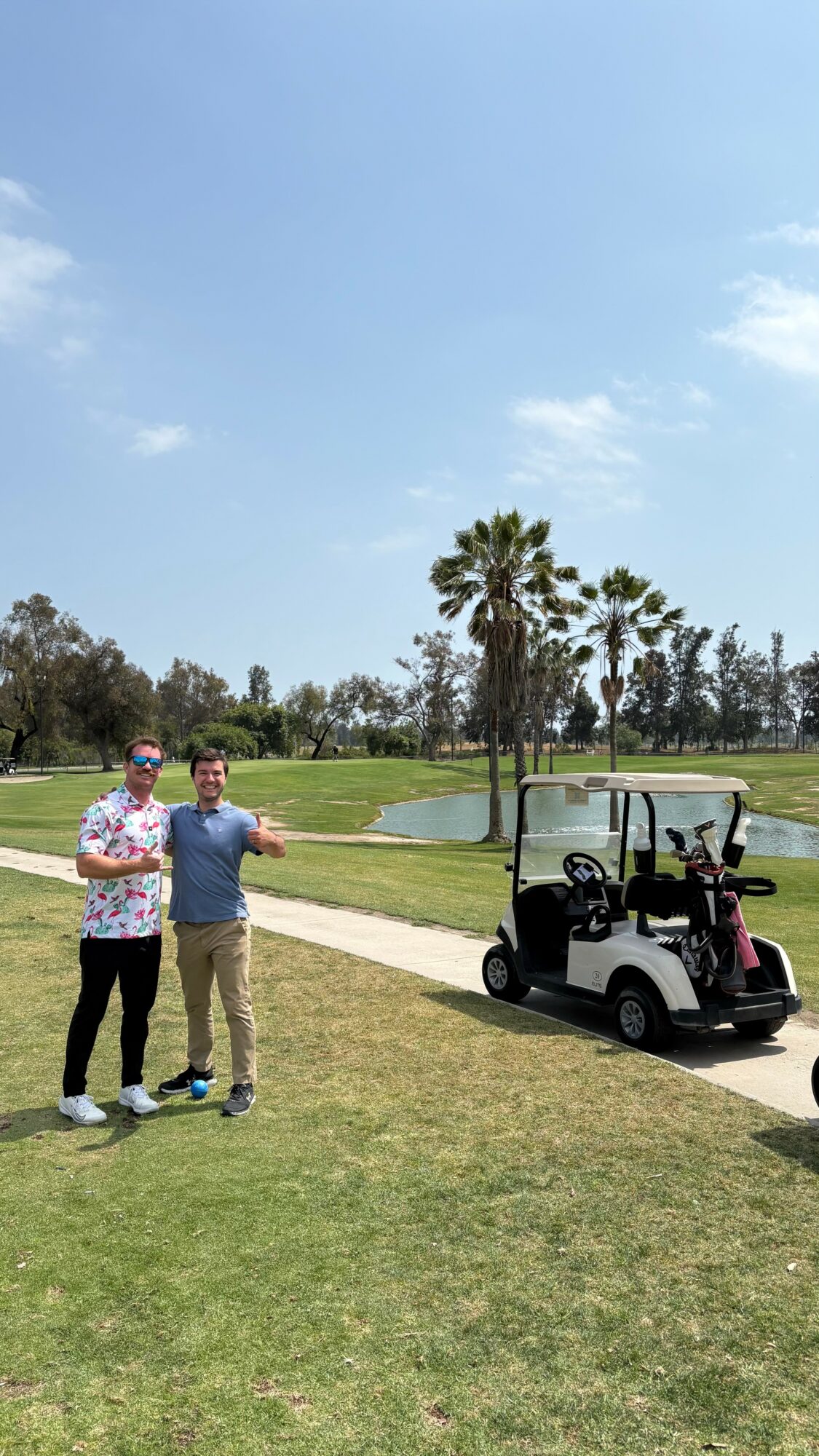 Two people stand on grass near a golf cart, with palm trees and a clear blue sky in the background.
