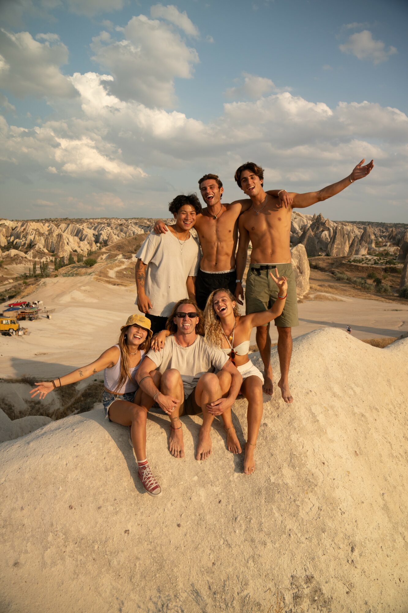 Group of six people posing outdoors on a sunny day, some standing and some sitting, with a rocky landscape and blue sky in the background.