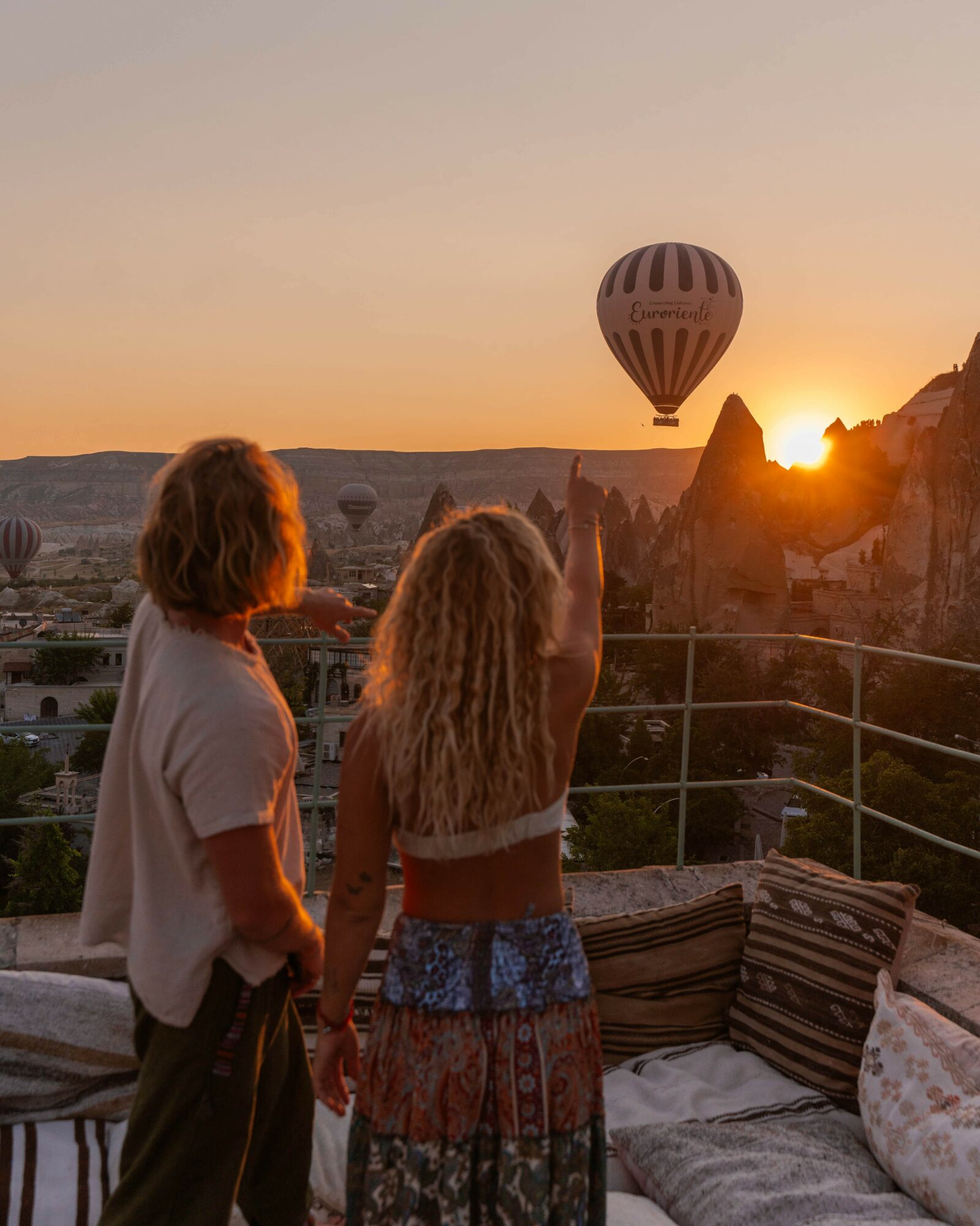 Two children watch a hot air balloon at sunset, with a landscape of rock formations and a railing in the foreground.