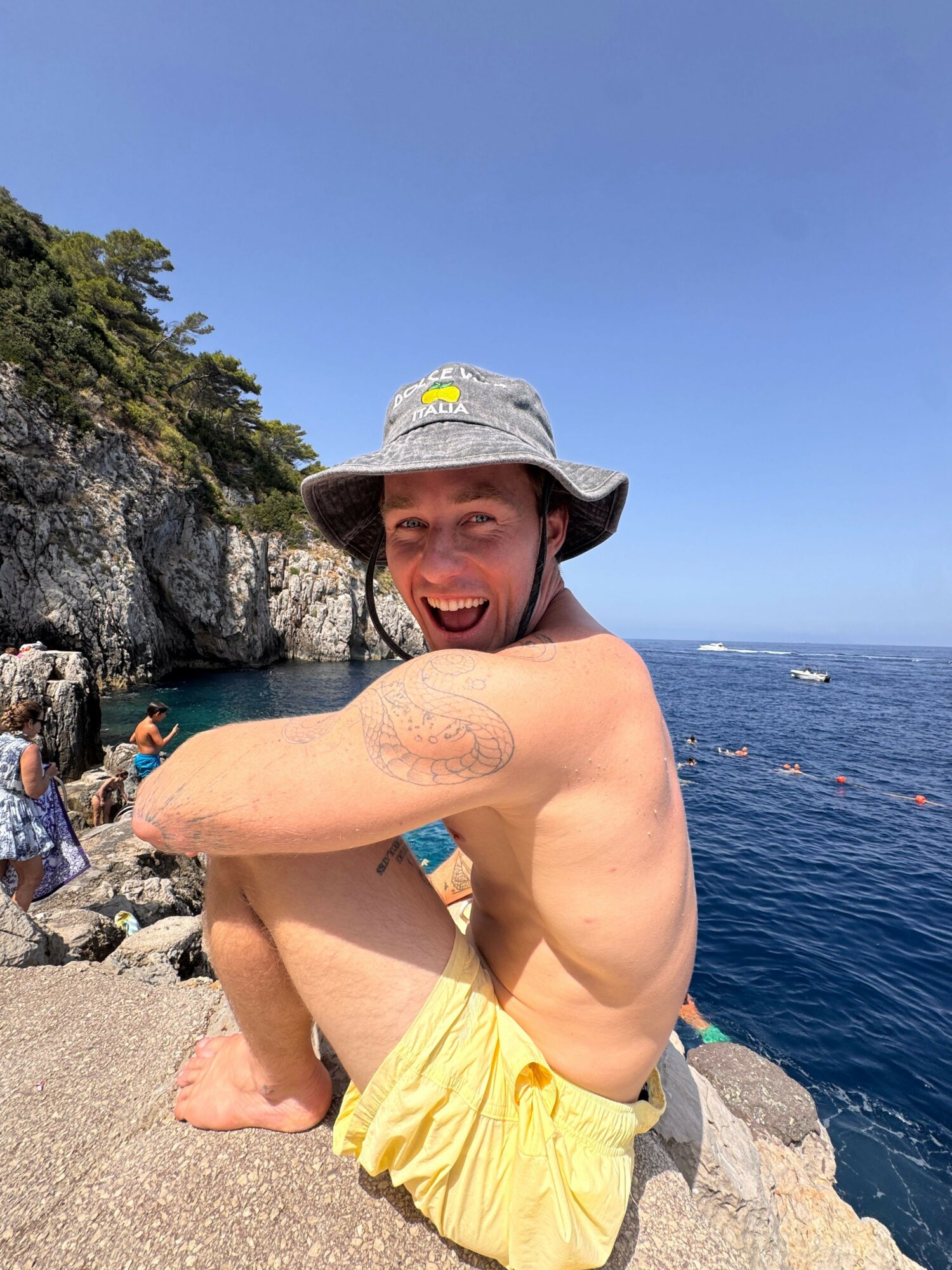 Person sitting on rocks near water, wearing a hat and yellow shorts, smiling, with a clear blue sky and trees in background.