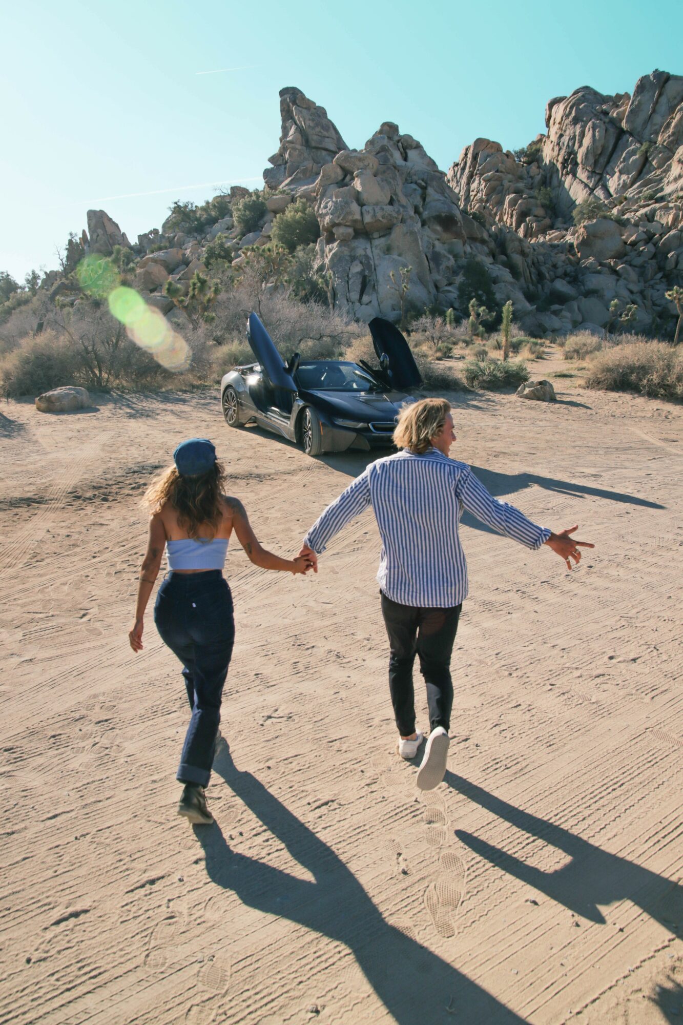Two people walk hand in hand on a sandy desert with rocky mountains in the background, casting long shadows.