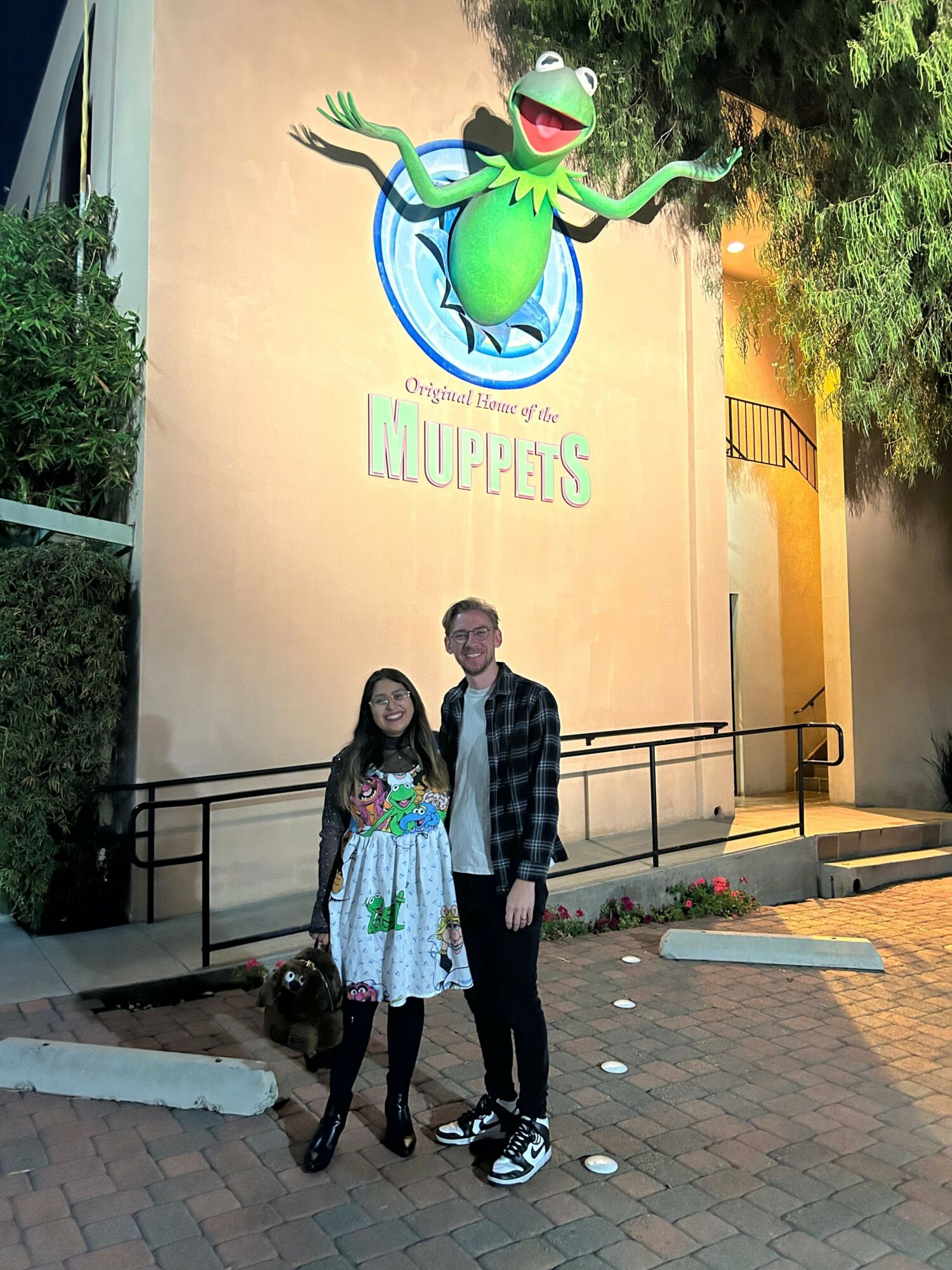 Three people standing in front of a wall with a large cartoon lizard and the word 'MOVIES' on it, outdoors at dusk.