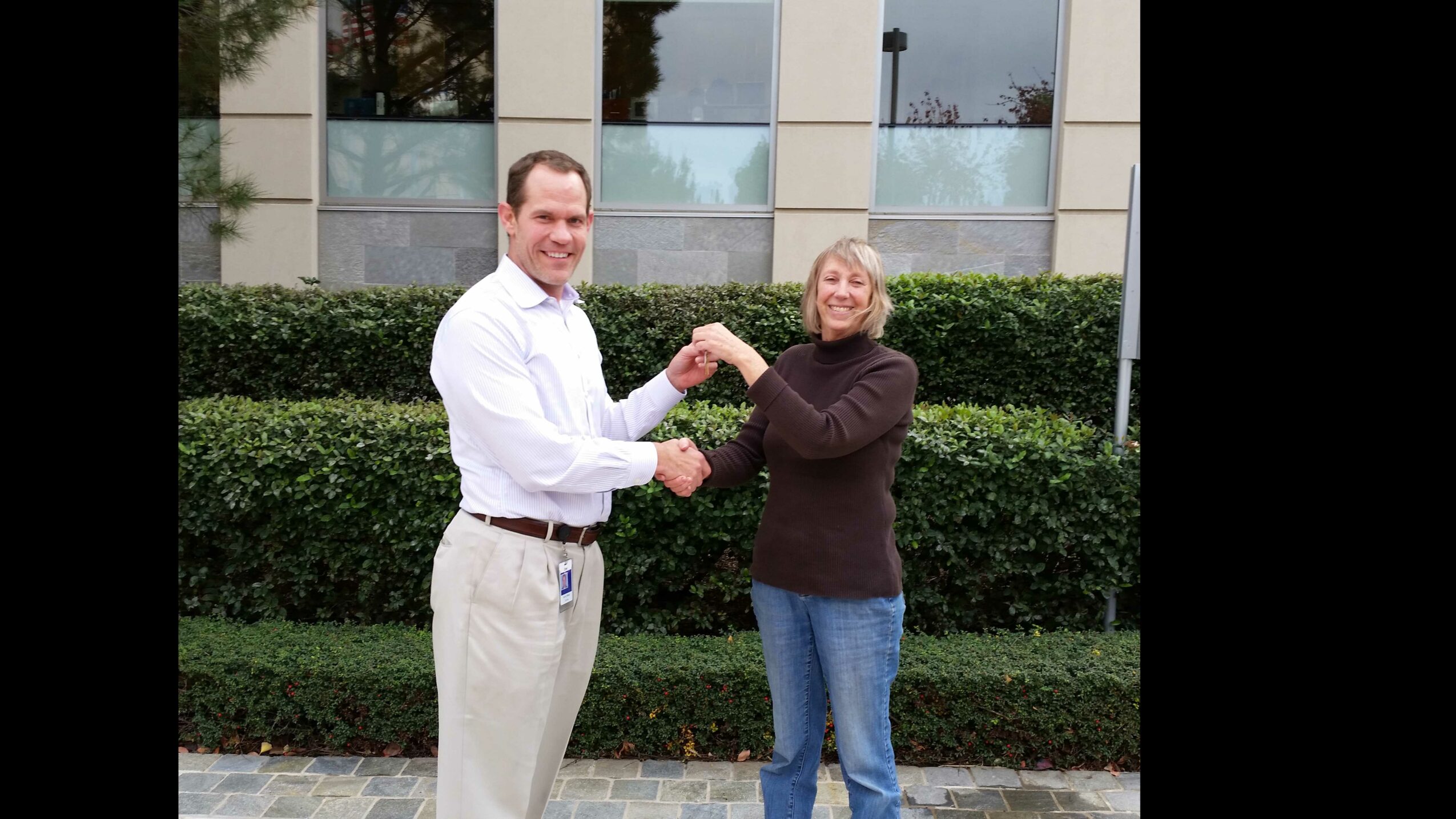 Two people exchanging a handshake outdoors in front of a hedge and building, smiling at each other.