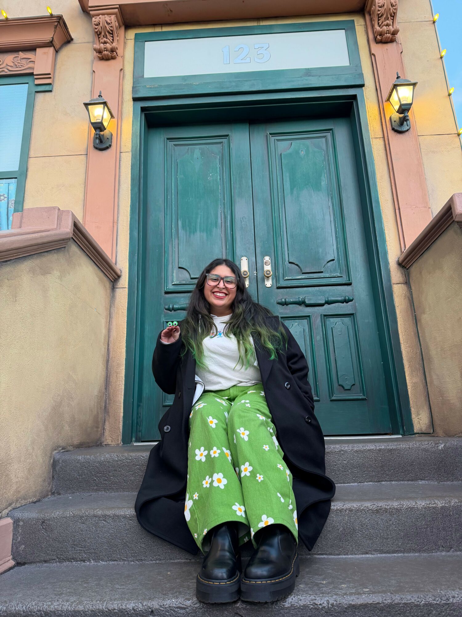 Person sitting on steps in front of a green door, wearing a black coat and green dress with white flowers, smiling.