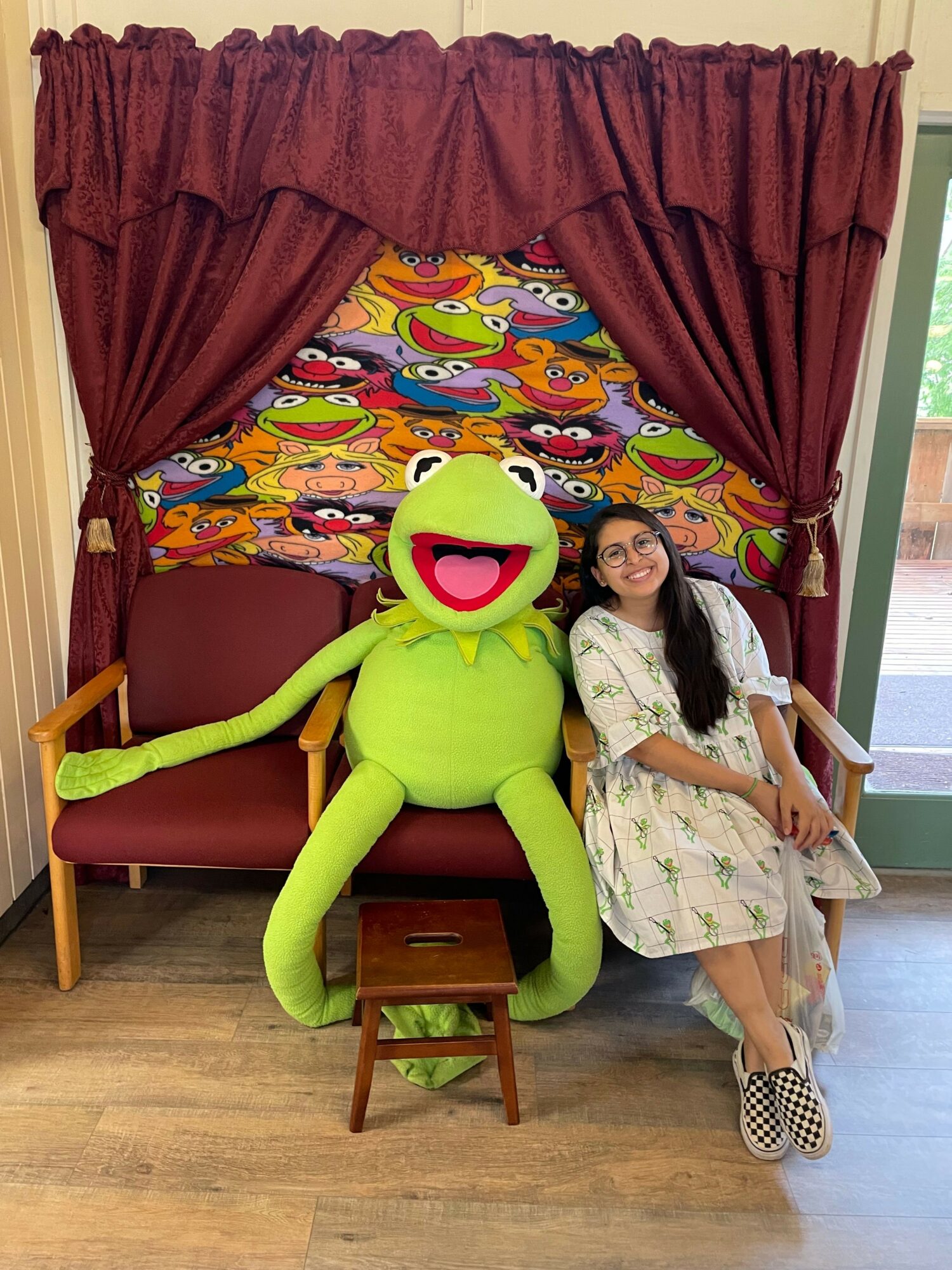 Girl lying on colorful bed with a large green frog plush toy, in a room with wooden floor and red curtains.